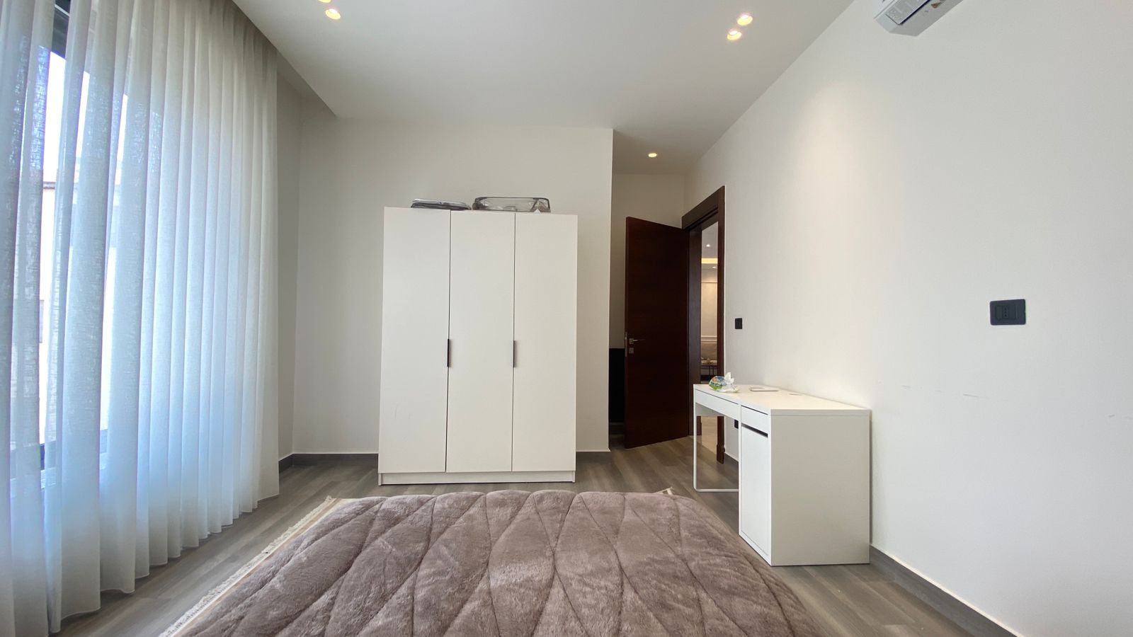 d7768c3d-93ed-4d4e-8b2d-61daf16cc73c Minimalist bedroom with a white wardrobe and a white desk against the right wall, a soft gray bedspread in the foreground, and sheer white curtains along a large window on the left; a dark wood door is open toward a dimly lit hallway.