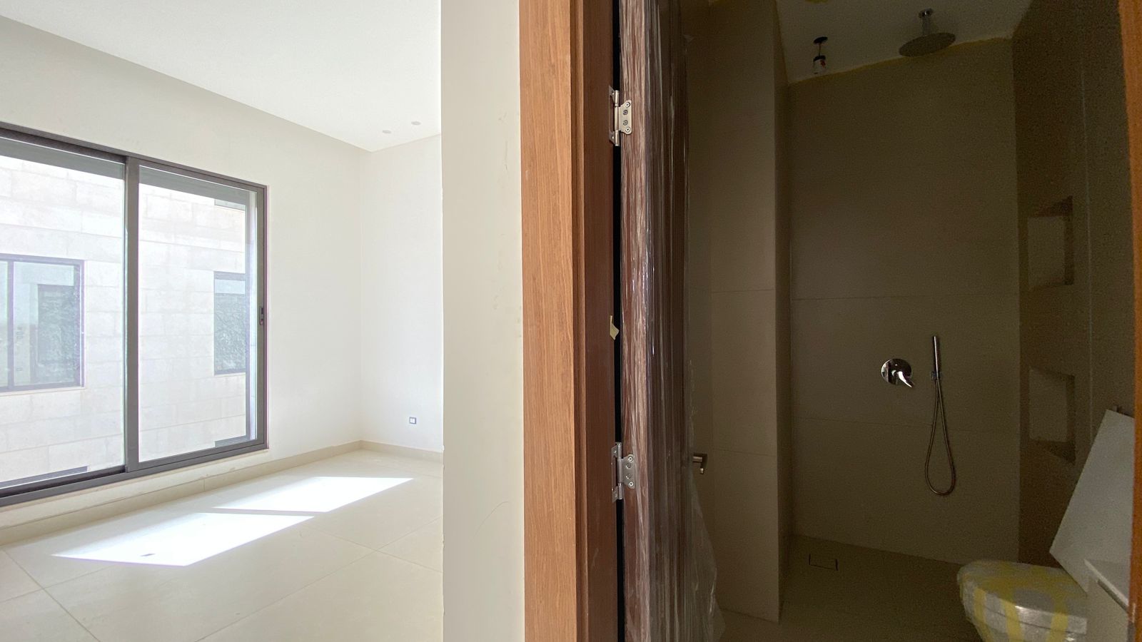 d7ae5f92-a79c-4807-9de2-7a55ea995f20 Bright white room with a large sliding window, sunlight on the tiled floor, and an open door to a dark adjacent bathroom with a shower fixture.