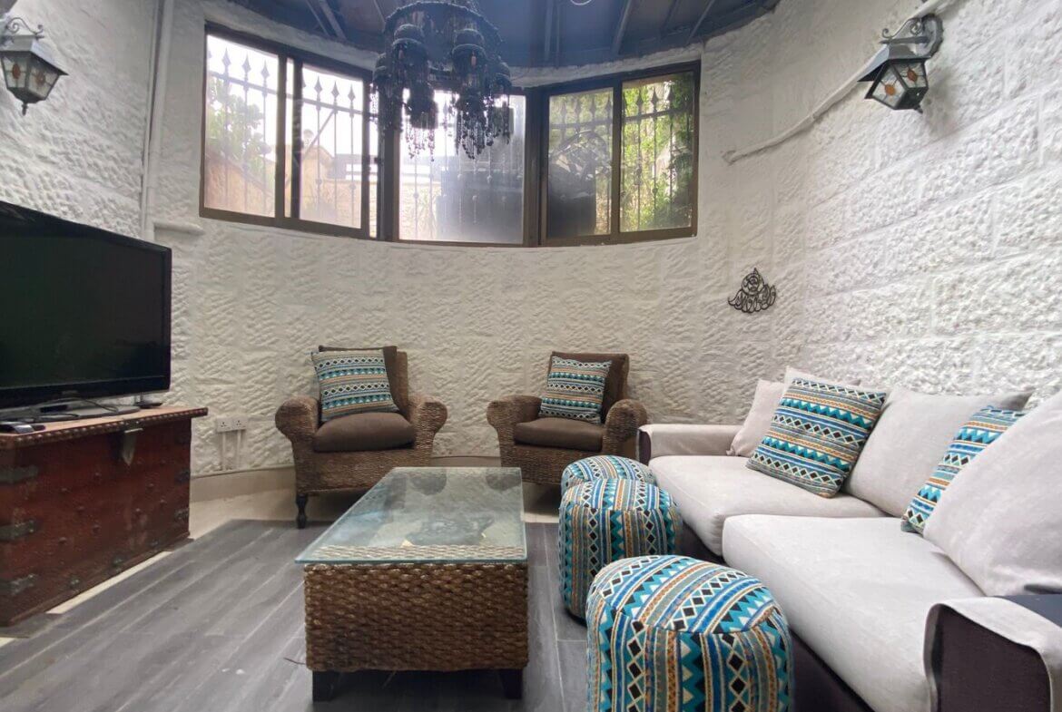 d875f150-cdab-4852-b8b1-9c2420d2f2c6 Cozy living room with white textured walls, beige sofa and wicker chairs, blue geometric cushions, glass-topped coffee table, and a TV on a vintage trunk.