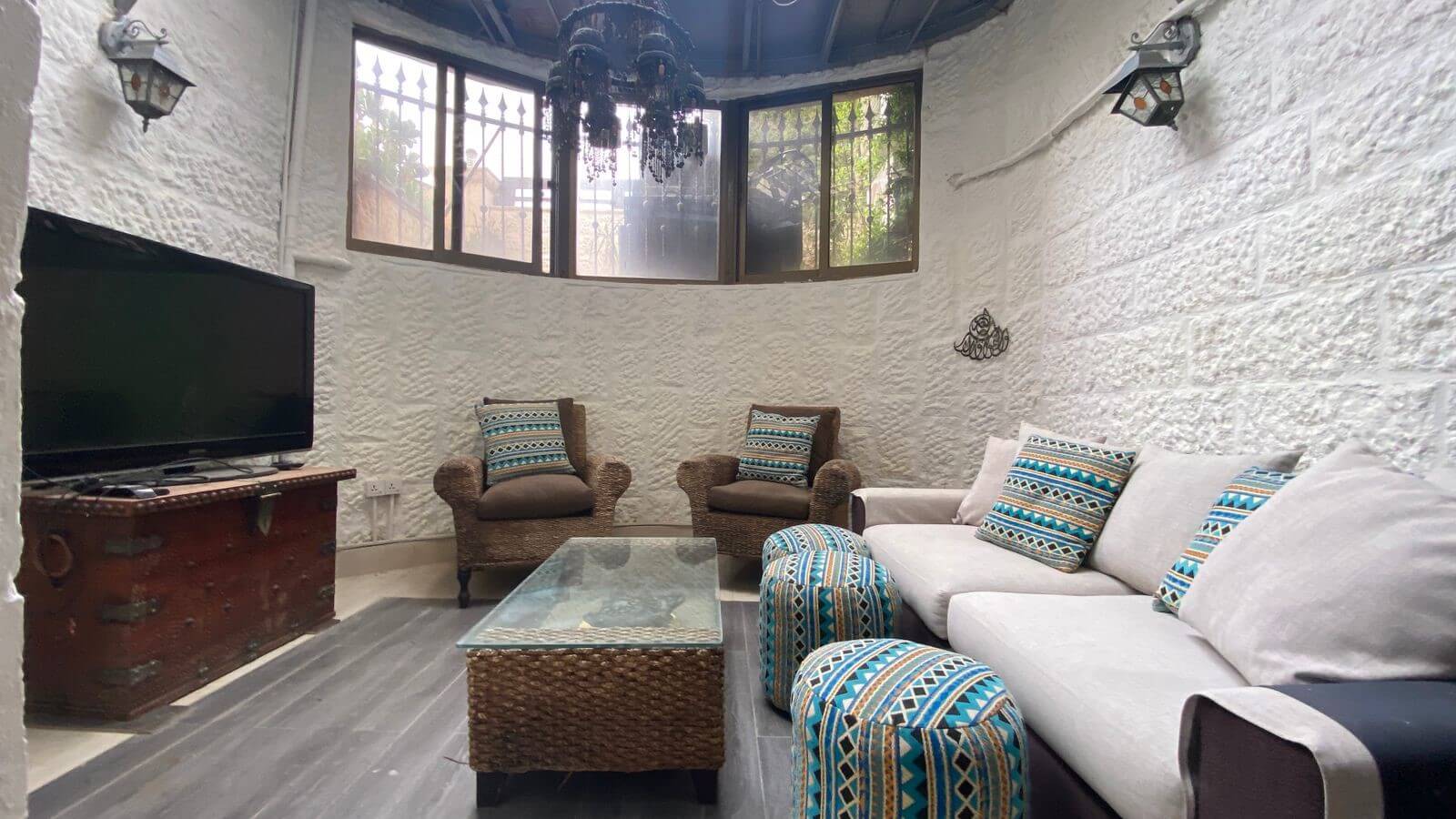 d875f150-cdab-4852-b8b1-9c2420d2f2c6 Cozy living room with white textured walls, beige sofa and wicker chairs, blue geometric cushions, glass-topped coffee table, and a TV on a vintage trunk.