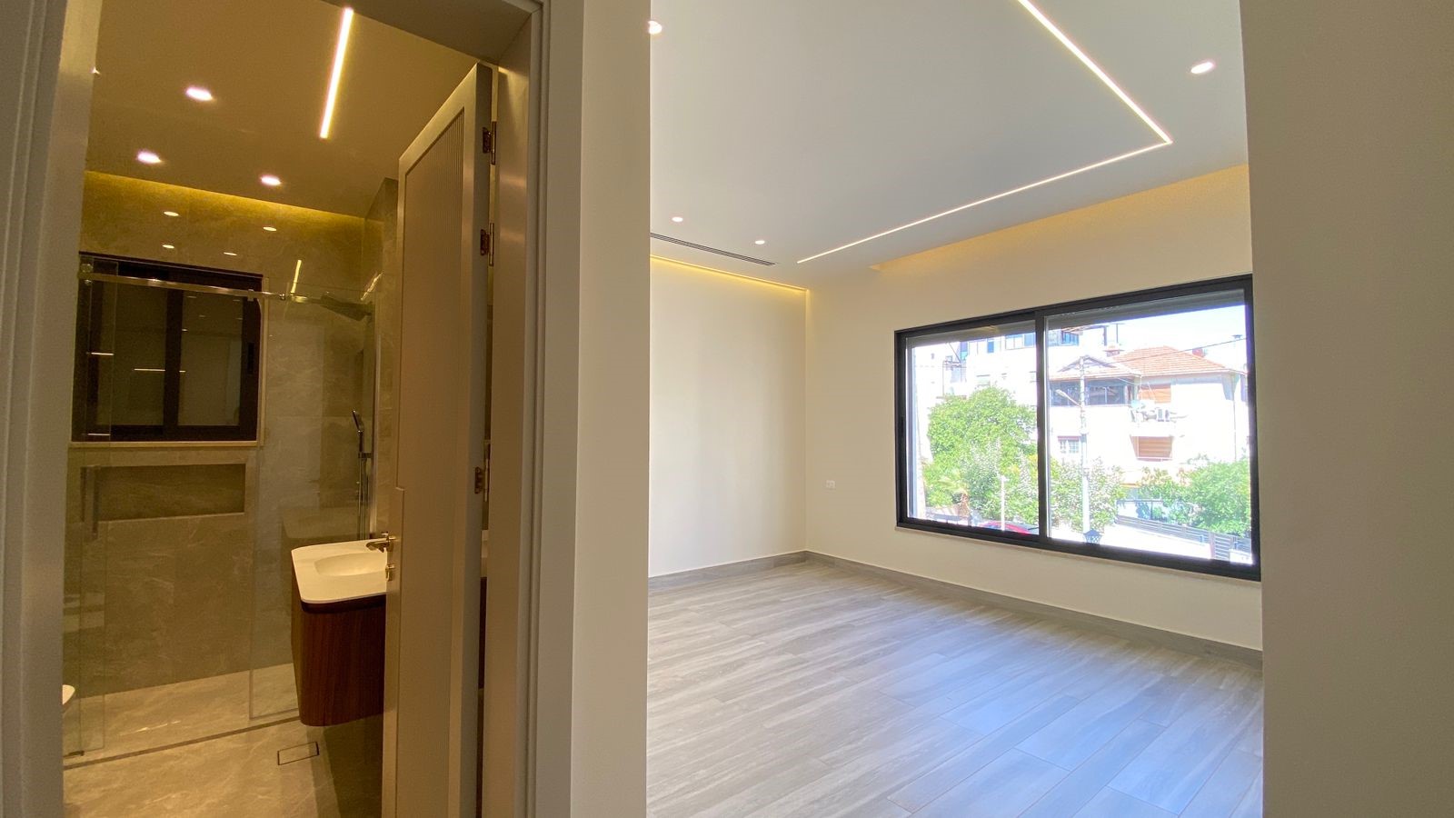 d8df25f1-c21f-4aee-881c-b61ac0980dc0 Empty modern room with large window, light gray wood floor, and ceiling LED lines; bathroom visible through an open doorway on the left.