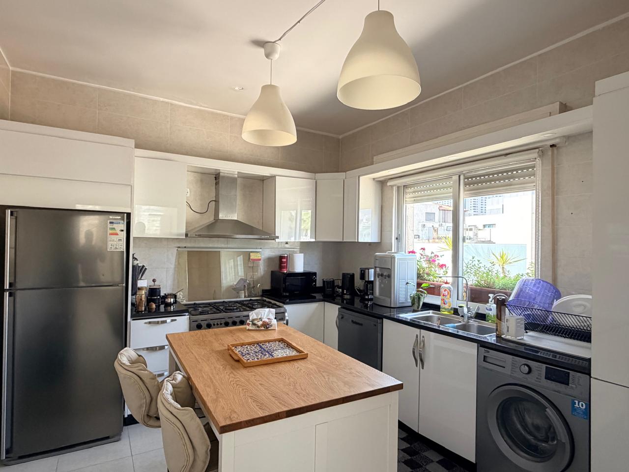 d9019ae0-0b95-4118-b45a-653854ce52fa Bright modern kitchen with island, white cabinets, and stainless steel fridge and oven. Window over sink with dish rack nearby.