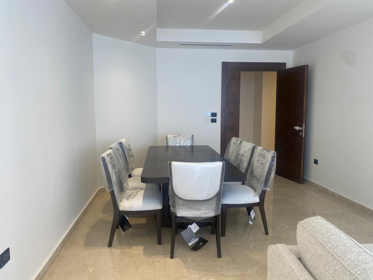 Modern dining room with a dark wooden table and six upholstered chairs still wrapped in protective plastic.