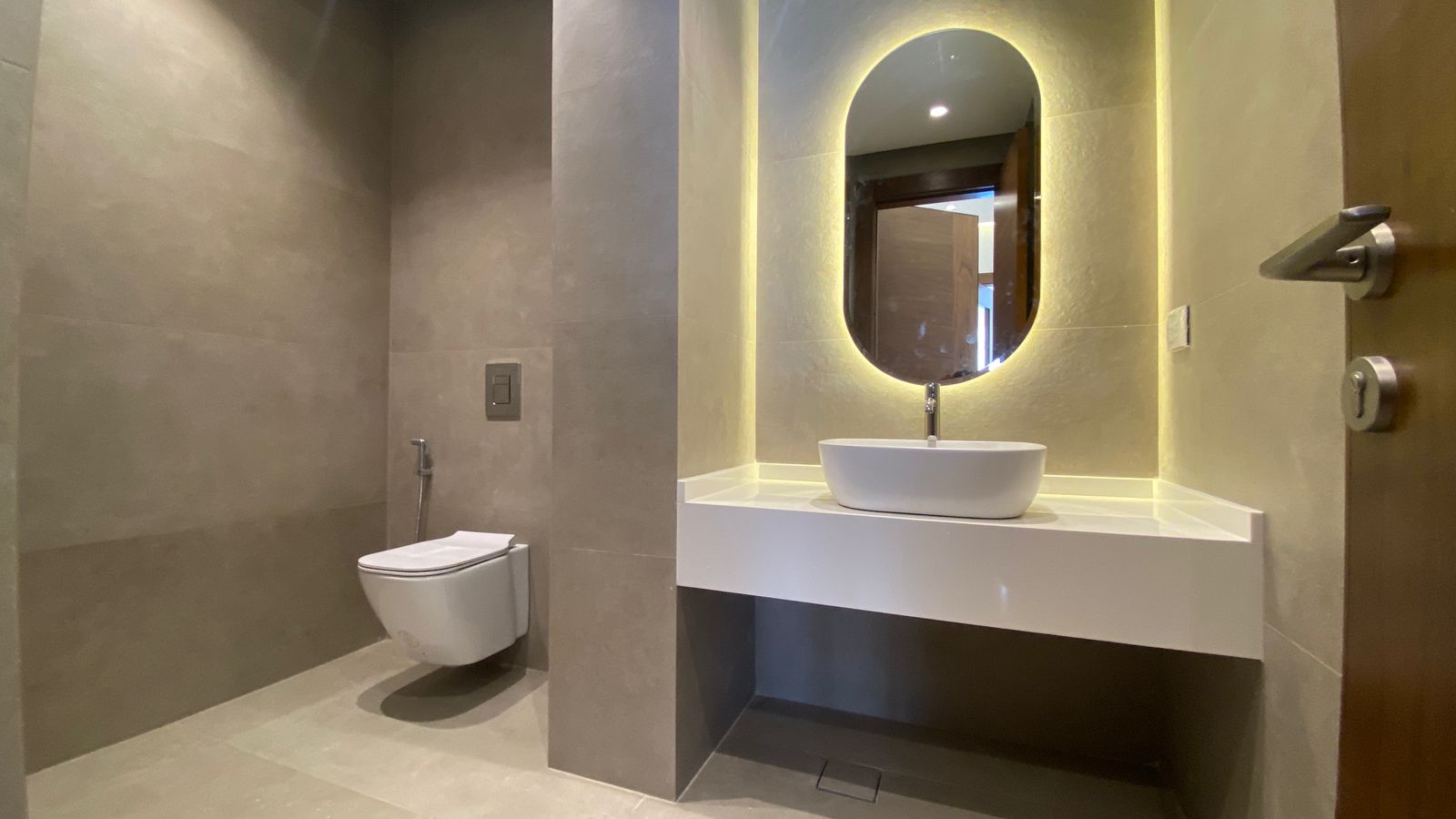 db426a66-46df-429f-b084-dd7b9dcca571 Contemporary bathroom with a wall-mounted toilet, a handheld bidet spray, and a white floating sink beneath a backlit oval mirror. The beige tile walls and warm lighting create a modern, minimalist feel.