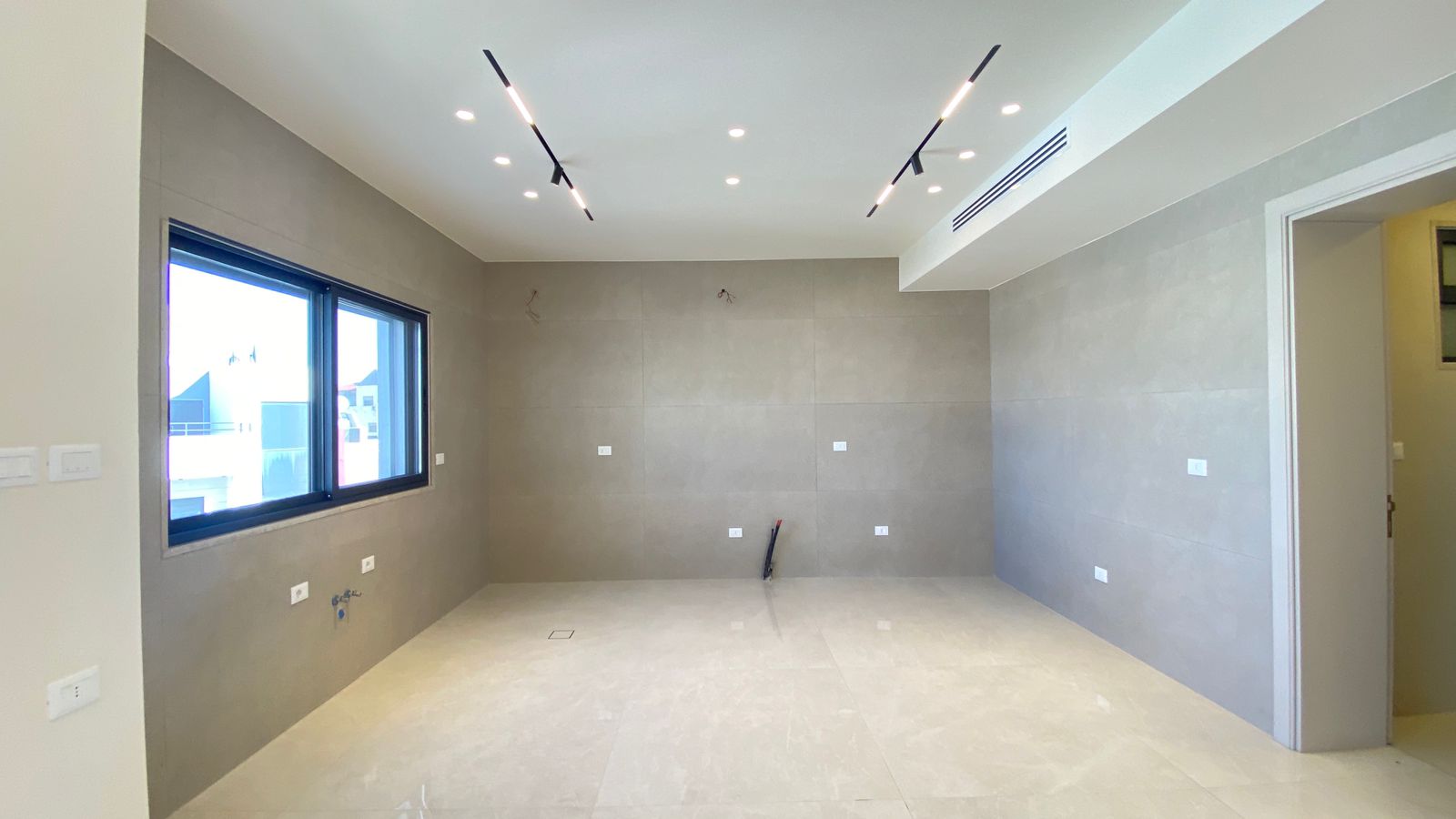 dbc4522a-5e53-47c2-a41e-d246fffa9f08 Unfurnished modern room with gray tiled walls, a large window, and track lighting; plumbing and outlets visible, suggesting a kitchen under construction.