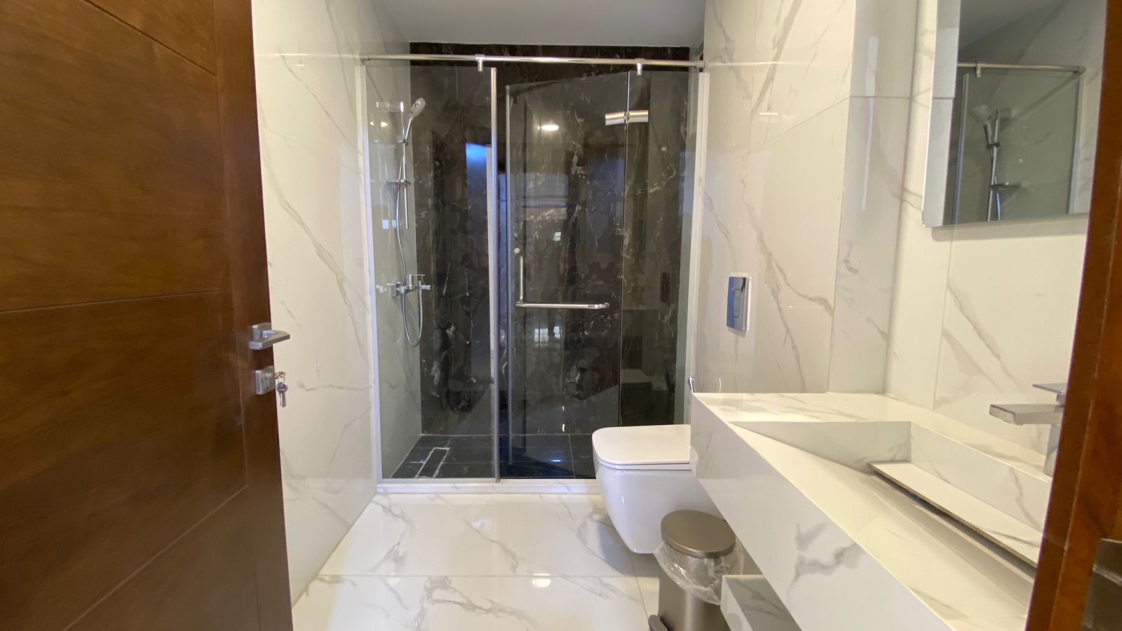 dc039857-8bfe-4f6d-a631-571b10e6b115 Modern bathroom with marble walls and floor, glass shower enclosure, white toilet, and white countertop sink area.