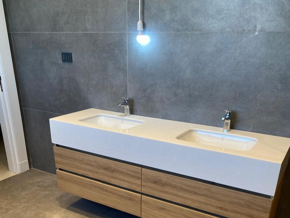 dc40eddf-bc1b-4edb-9cb0-e0ed910873e0 Double bathroom sink vanity with white countertop and wood-tone drawers against gray tiled wall, single hanging bulb light above