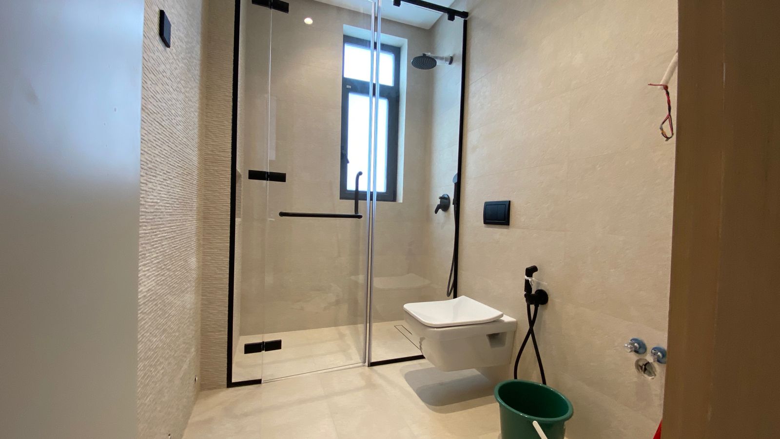 dc967308-10e5-4096-a0d2-0239247614b9 Glass-walled shower area with black hardware and a wall-hung toilet in a beige-tiled bathroom, back window visible.