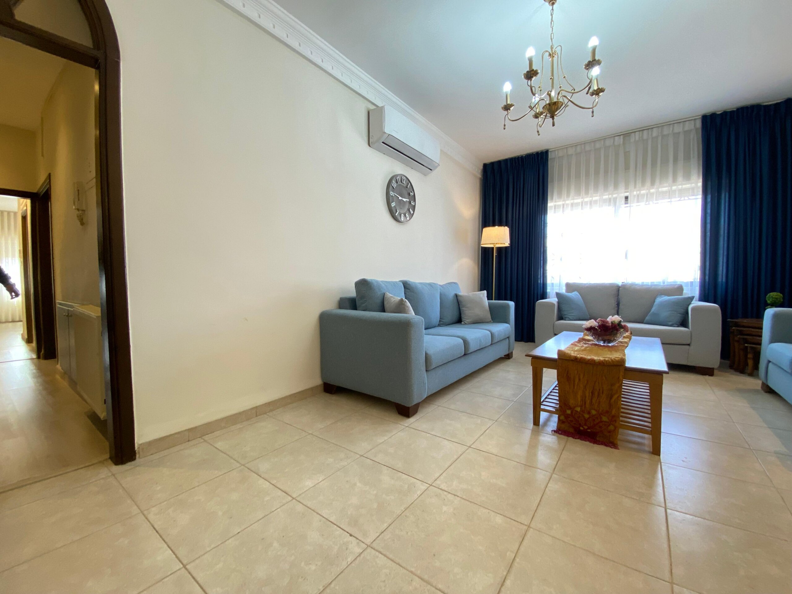 dcc726af-bcd8-4271-b54f-c45cf9f80d82 Living room with beige walls, blue sofas, wooden coffee table, and flowing dark blue curtains by large windows with sheer drapes. A chandelier hangs from the ceiling.
