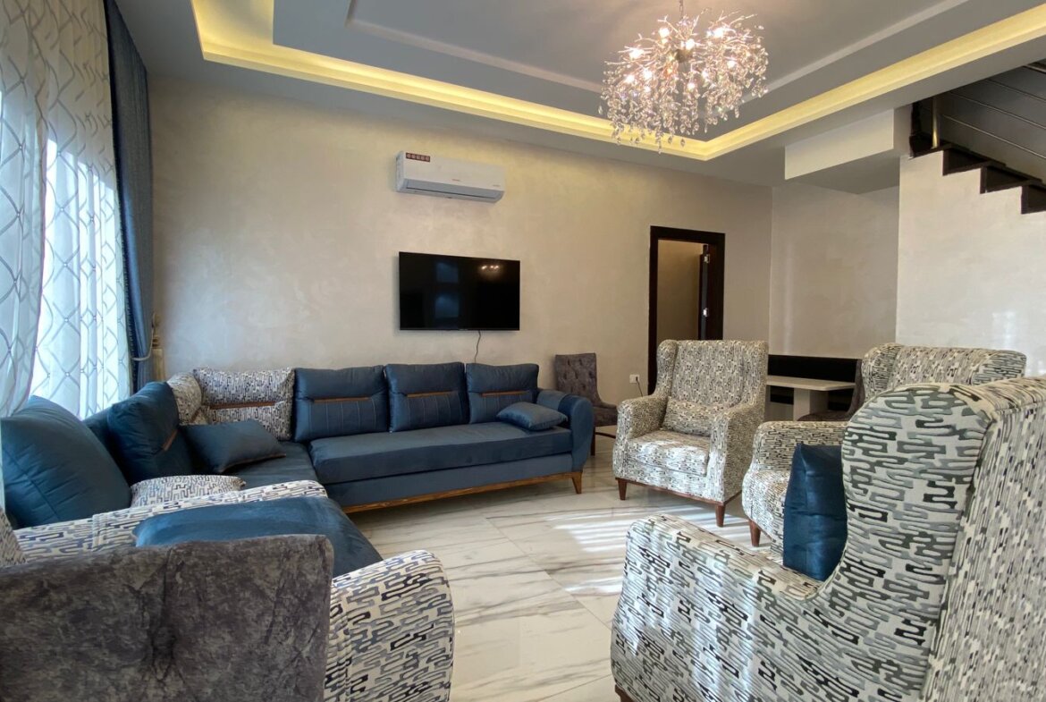 Modern living room with blue sectional sofa, patterned armchairs, and a wall-mounted TV under an LED ceiling cove light.