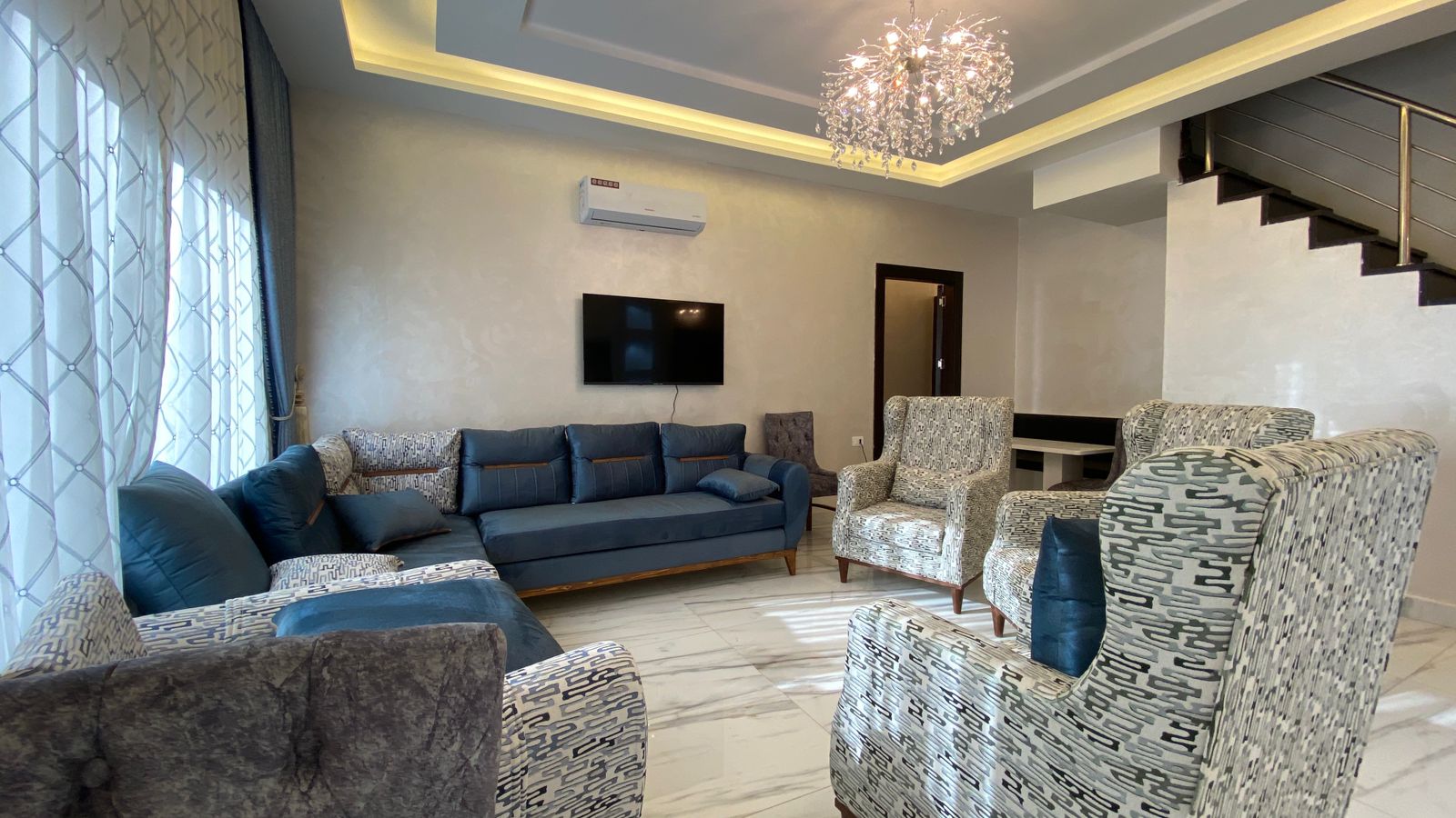 Modern living room with blue sectional sofa, patterned armchairs, and a wall-mounted TV under an LED ceiling cove light.