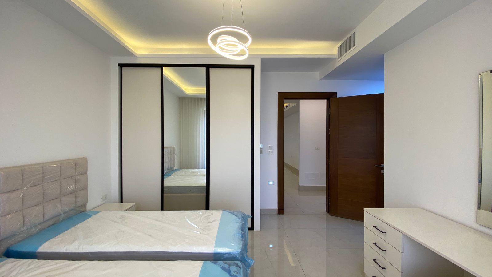 de366efd-6f7f-4e6f-b63c-33b78ea133bf Modern bedroom with white furniture: two twin beds in plastic, a mirrored sliding-door wardrobe, and a white desk. Open wooden door leads to a hallway.