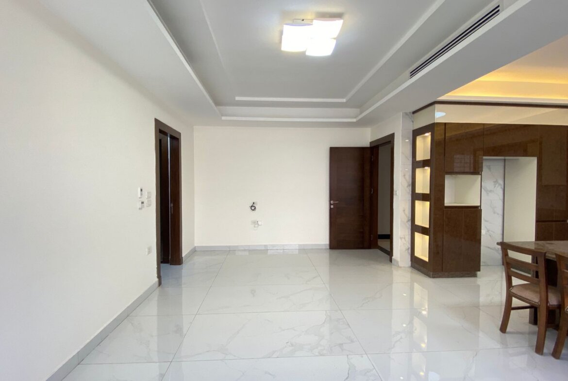 Spacious open-concept room with white walls, glossy marble floor, and dark wooden kitchen cabinets on the right; a wooden dining table and chairs in the foreground.
