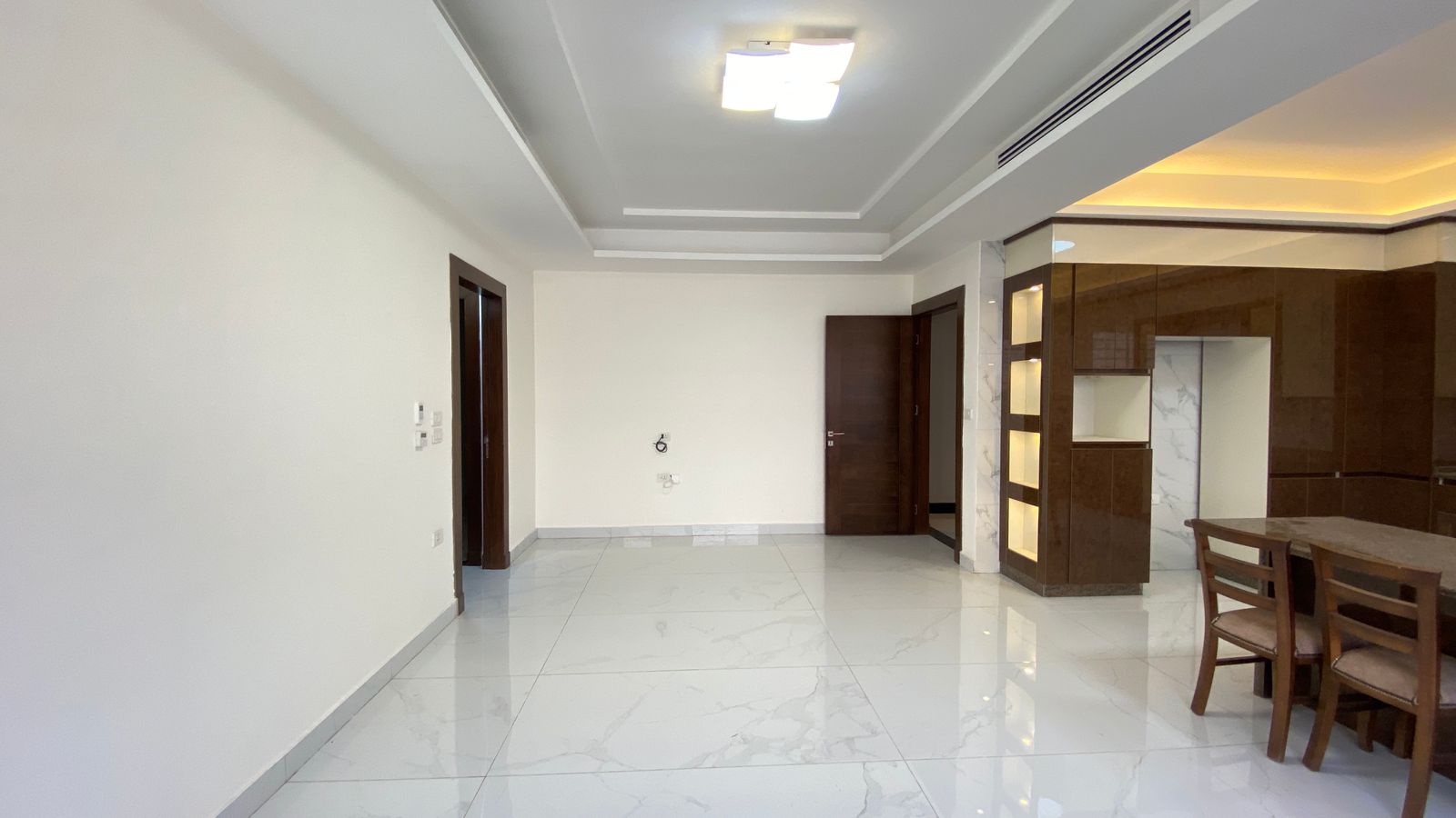 de369f0e-9f8e-411e-9e9e-418b37303155 Spacious open-concept room with white walls, glossy marble floor, and dark wooden kitchen cabinets on the right; a wooden dining table and chairs in the foreground.