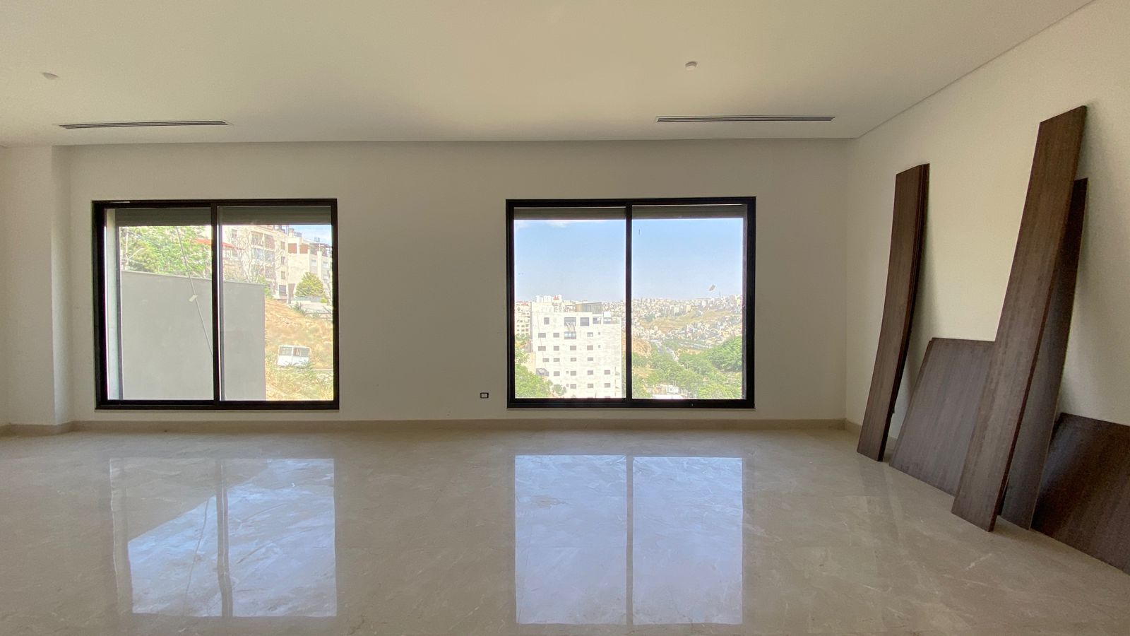 de48cf5c-6d9a-4f1d-b2f9-7413dc9398d9 Empty bright living area with two large windows showing a city view; several wooden planks lean against the right wall.