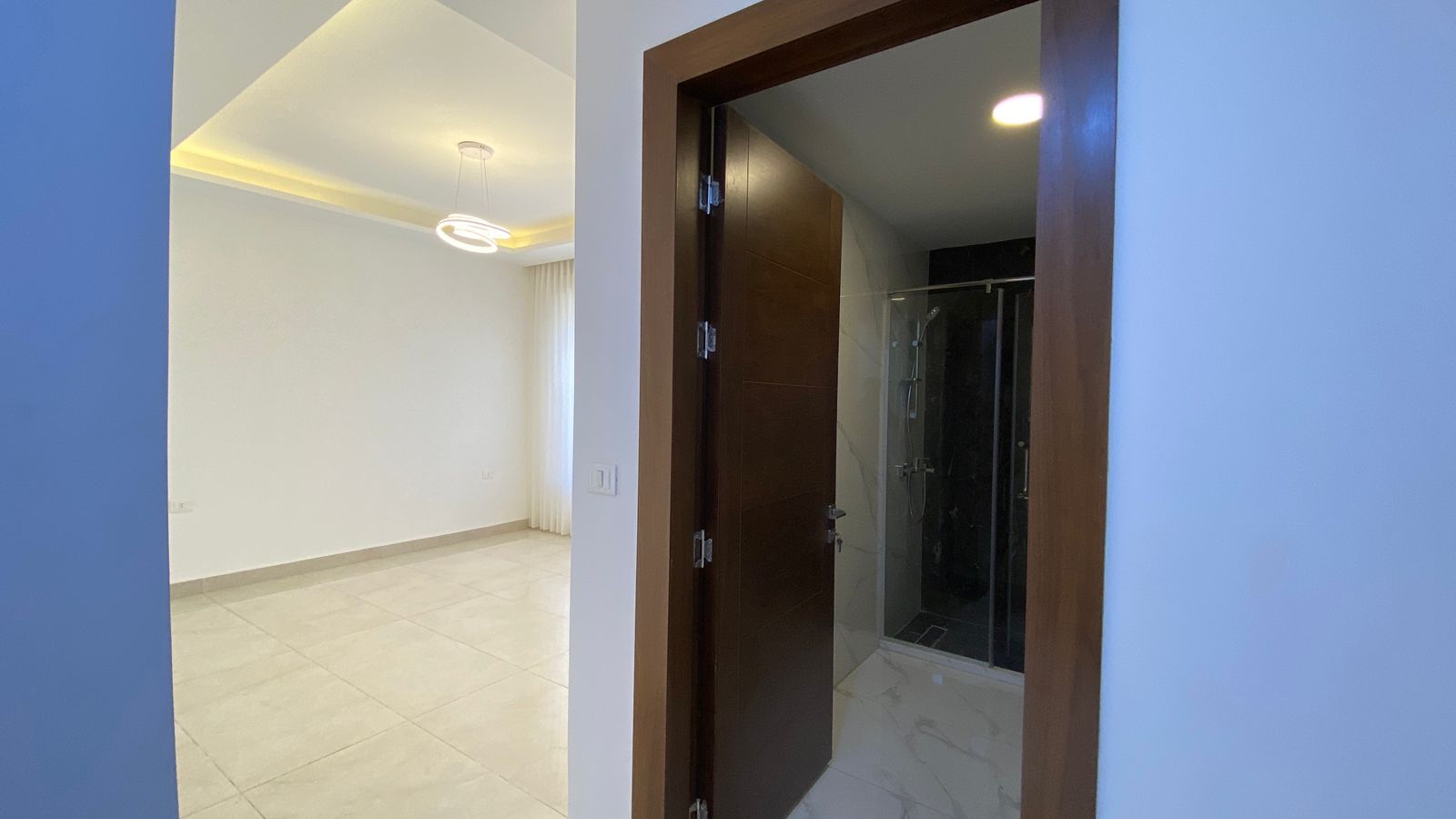 df025488-0b0a-4556-8626-d279b9876a09 Bright modern living area with tiled floor and large window covered by sheer curtains; open doorway to a bathroom on the right.