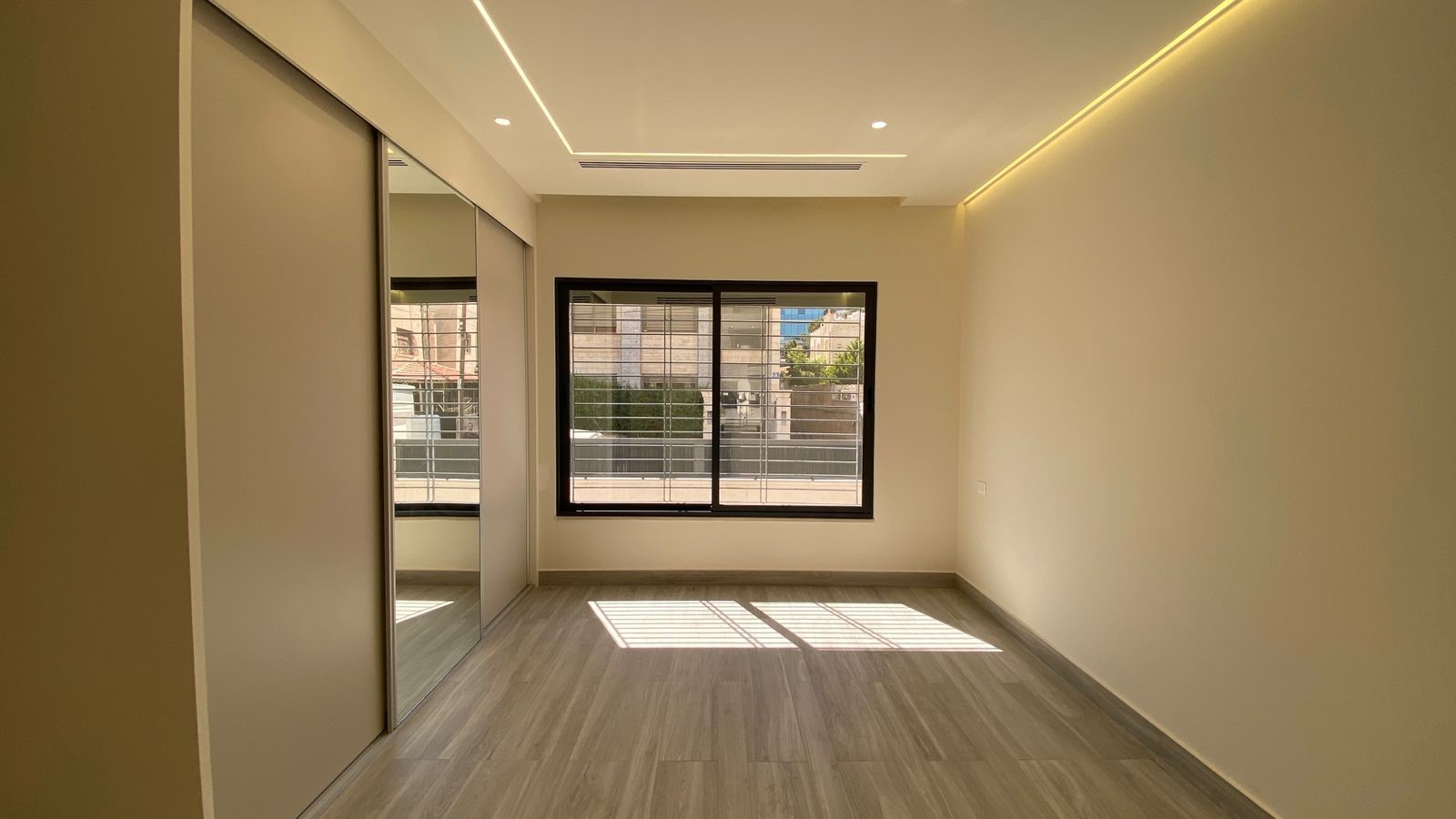 df14e634-8970-4170-83e2-25b639918785 Empty modern bedroom with a large window, mirrored sliding doors on the left, and warm ceiling lighting.
