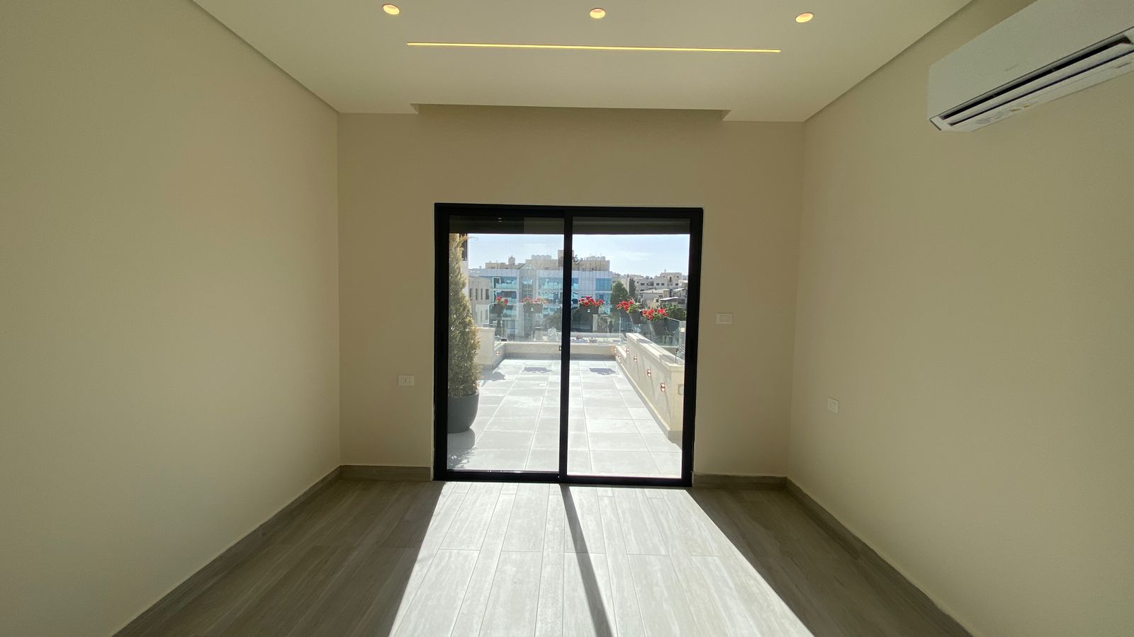 df25f9d6-9082-4da3-853e-8e7c47d19ae6 Empty modern room with a glass sliding door leading to a terrace; city buildings visible outside, light wood-look flooring, neutral walls, and ceiling lights.