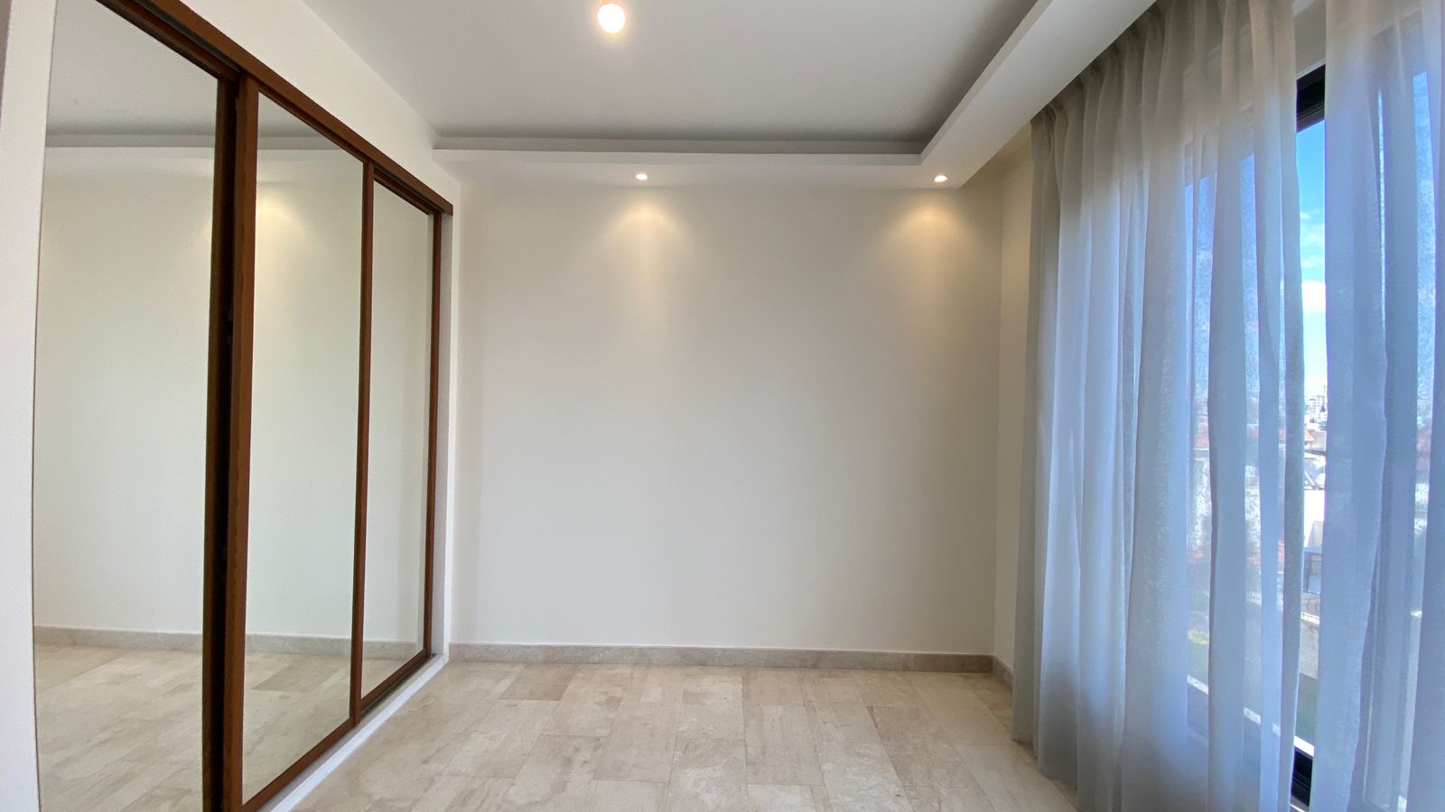 df3a33f4-d386-466e-ab14-7da47dfd8aa5 Empty modern room with mirrored sliding doors on the left, white walls, and sheer curtains over a large window on the right.