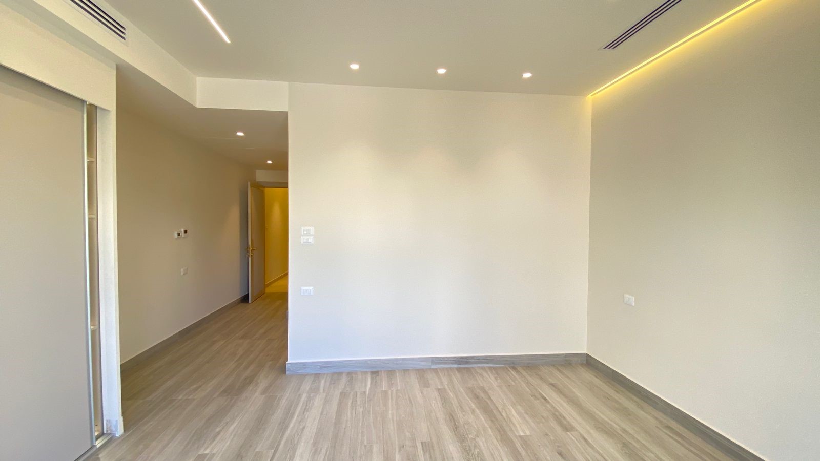 e0285173-a1fc-4d75-ac82-53cd9082d17a Empty modern living space with light wood flooring, white walls, recessed ceiling lights, and warm LED strip lighting along the right wall.
