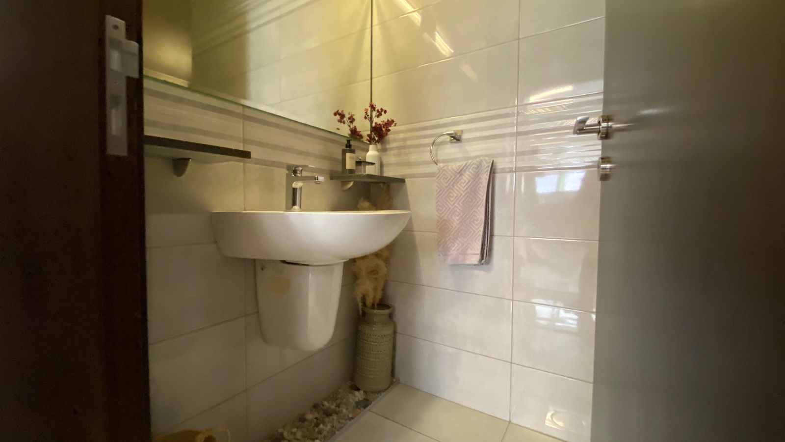 e03ddb3c-27a5-4c4b-8d20-f1d1866dd1f5 Small modern bathroom with wall tiles, white pedestal sink, towel ring, and a striped towel; decorative vase and shelf above sink; vase with dried flowers on the floor nearby.