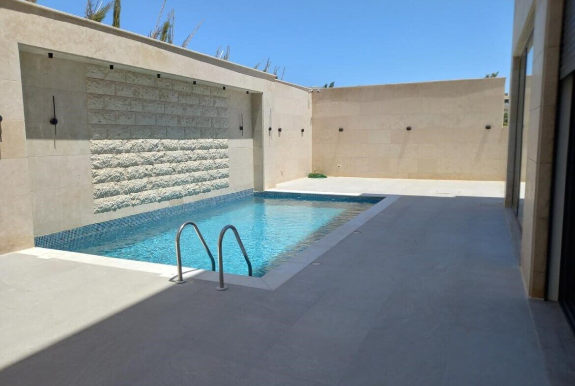 e0db4078-8aa9-451b-97af-1ab4b686acd5 Rectangular in-ground pool with a metal ladder, blue-tiled edge, and a beige stone wall enclosure featuring a built‑in waterfall.