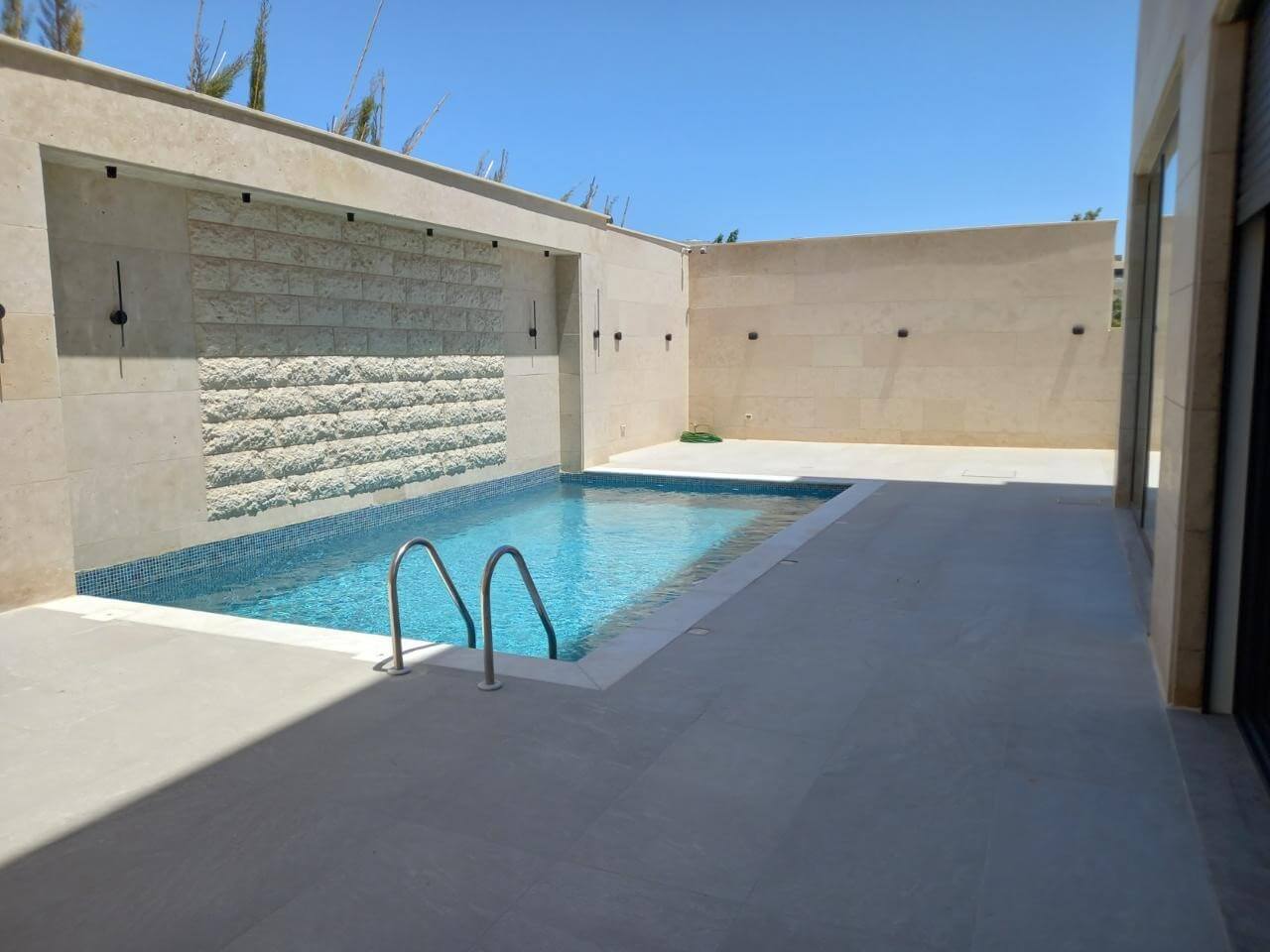 e0db4078-8aa9-451b-97af-1ab4b686acd5 Rectangular in-ground pool with a metal ladder, blue-tiled edge, and a beige stone wall enclosure featuring a built‑in waterfall.