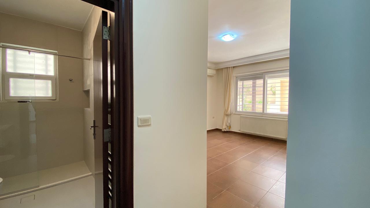 e19020fd-bb57-41a6-947e-1170b42e306a View into a modern bathroom with a glass shower door on the left; hallway opens to a sunlit living room with a large window and radiator.
