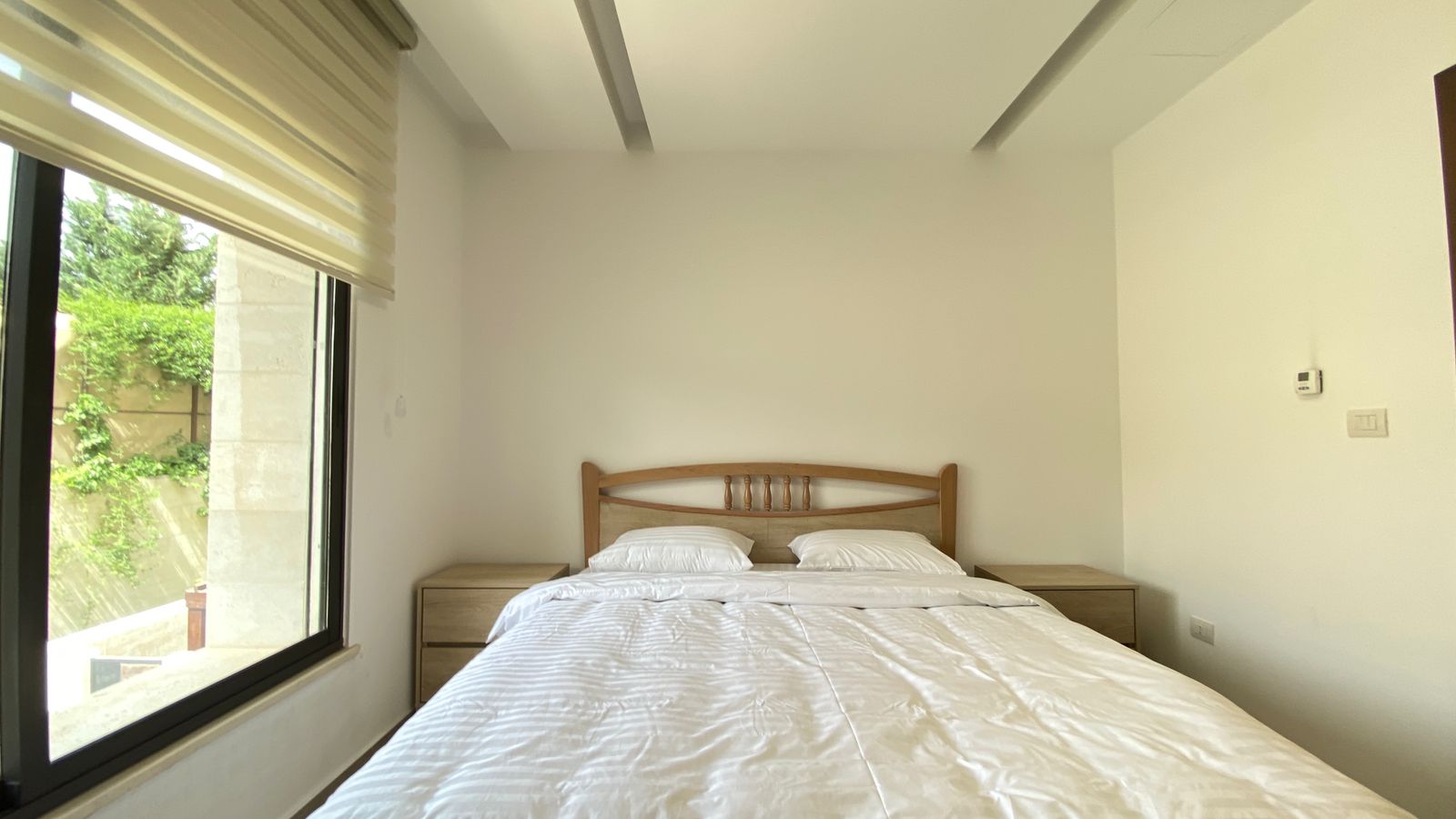 e1969c76-b934-43aa-95ff-636092207366 Bedroom with a wooden headboard bed, white linens, and two matching nightstands in a bright, minimalist room with a large left window showing greenery outside