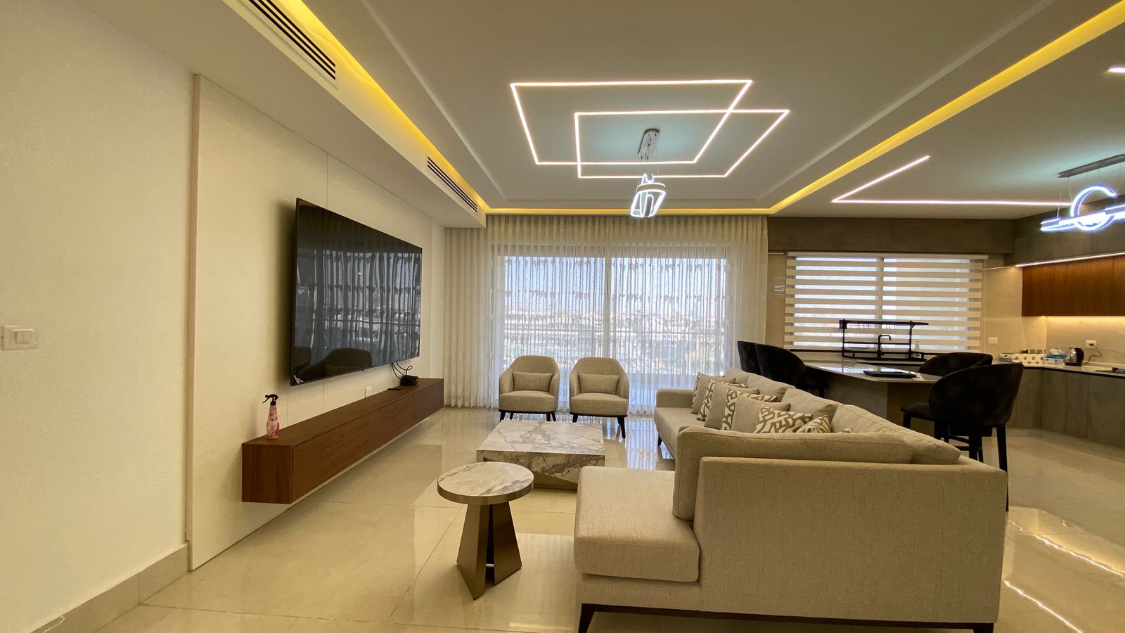 e2048ea3-7efe-44ea-ab56-61163f0d5ea1 Open-concept living room with a wall-mounted TV, low wooden media console, and beige seating facing a glass sliding door with sheer curtains.