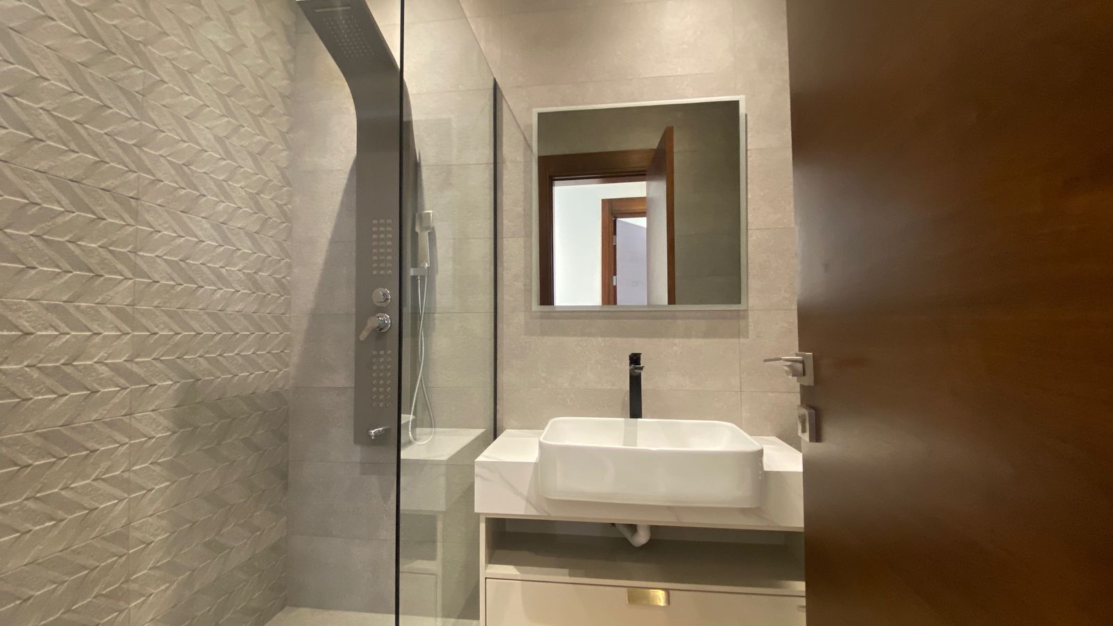 e2ec4fc6-e175-450a-b6c7-8451a1cf112b 1 Modern bathroom with a glass shower enclosure, textured chevron tile wall, white rectangular sink, and a square mirror above the vanity.