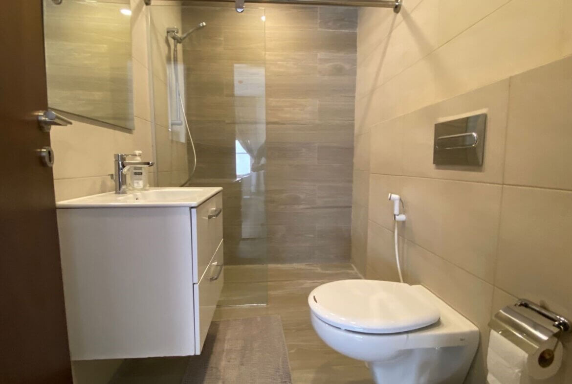 e302e7cc-d115-4b16-8062-fddfe7c37989 Small modern bathroom with white vanity, sink, and glass-enclosed shower seen from doorway; tiled walls and a wall-mounted toilet with a bidet sprayer nearby.