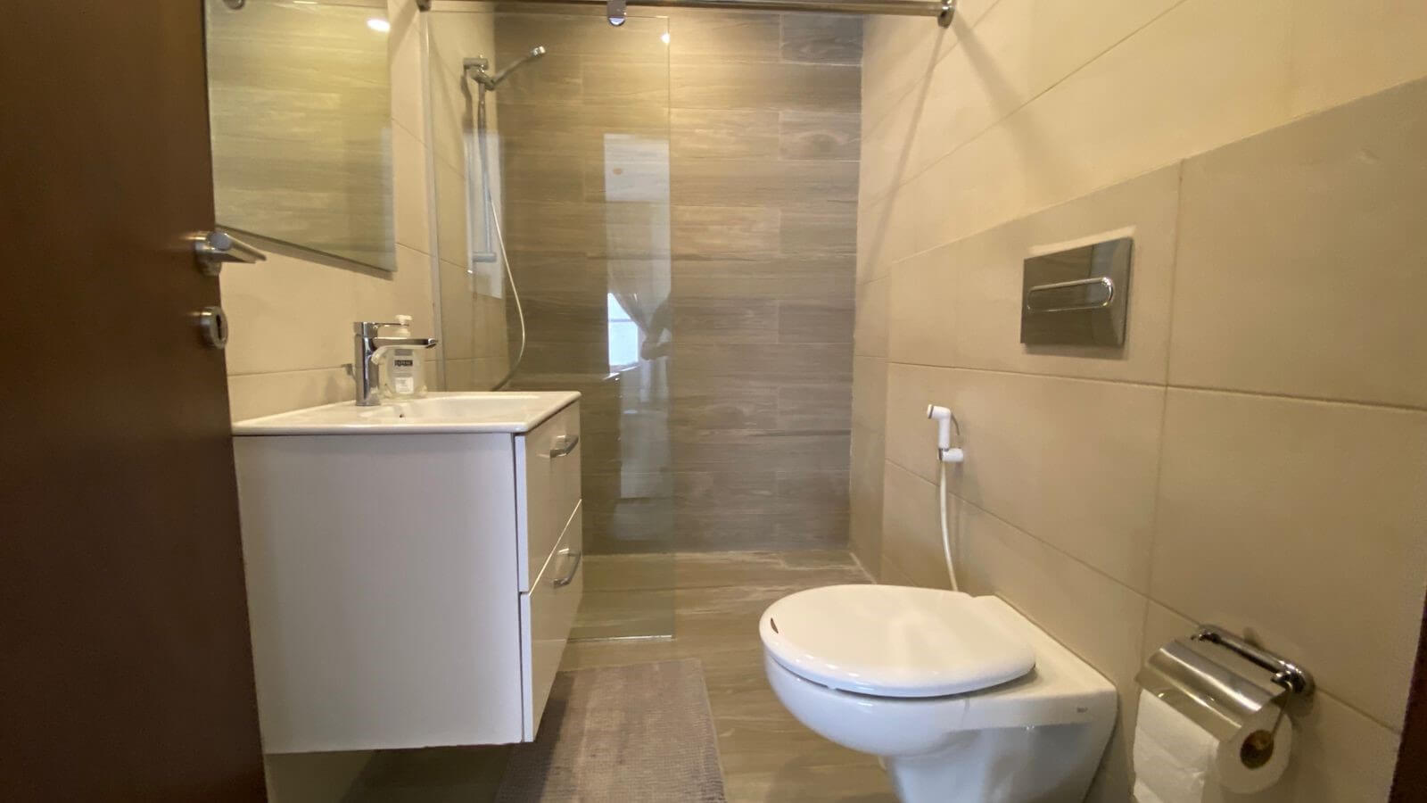 e302e7cc-d115-4b16-8062-fddfe7c37989 Small modern bathroom with white vanity, sink, and glass-enclosed shower seen from doorway; tiled walls and a wall-mounted toilet with a bidet sprayer nearby.