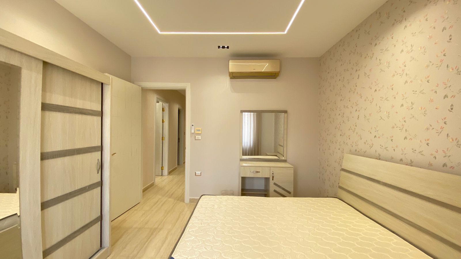 e31aa436-4727-4c95-a36a-1c63dc6a6f55 Bright bedroom with pale wood furniture, a queen bed, sliding wardrobe, and a vanity with mirror along a floral wallpapered wall.