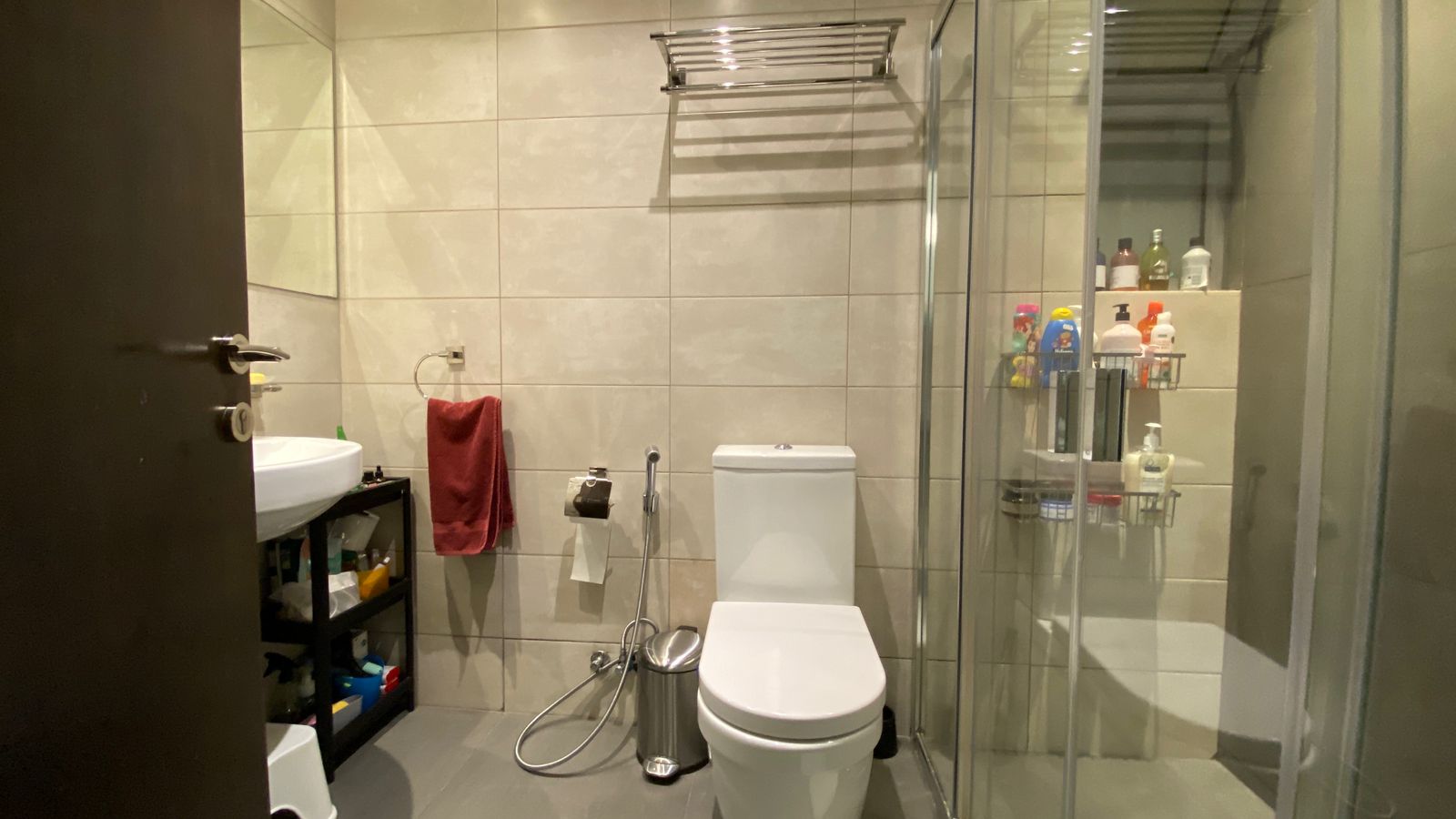 e31d7f63-56df-4be8-b5e4-34c9dea3f540 Contemporary bathroom with a white toilet, towel rack, bidet spray, and glass shower enclosure; beige tiled walls.