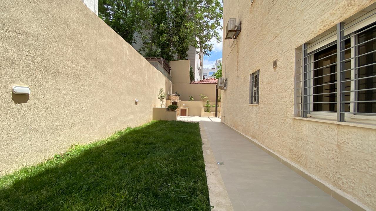 e3366405-fd64-430e-b8d6-ef460f1ea1a6 Narrow sunlit alley between beige stucco walls with a grassy strip on the left, a gray walkway on the right, and windows with bars along the building.