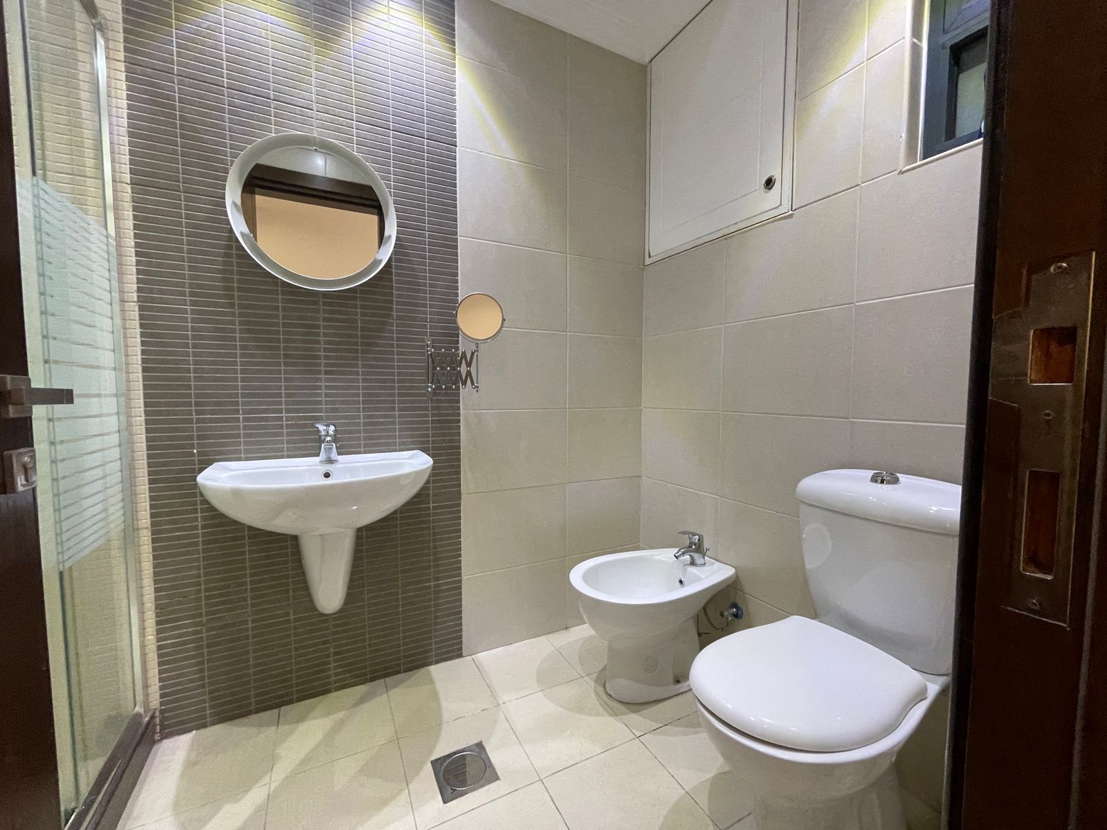 e3a1951d-0163-4146-a0c3-3d12dfa50993 Modern bathroom with white pedestal sink, round wall mirror, bidet, and toilet against beige walls and a glass shower door nearby