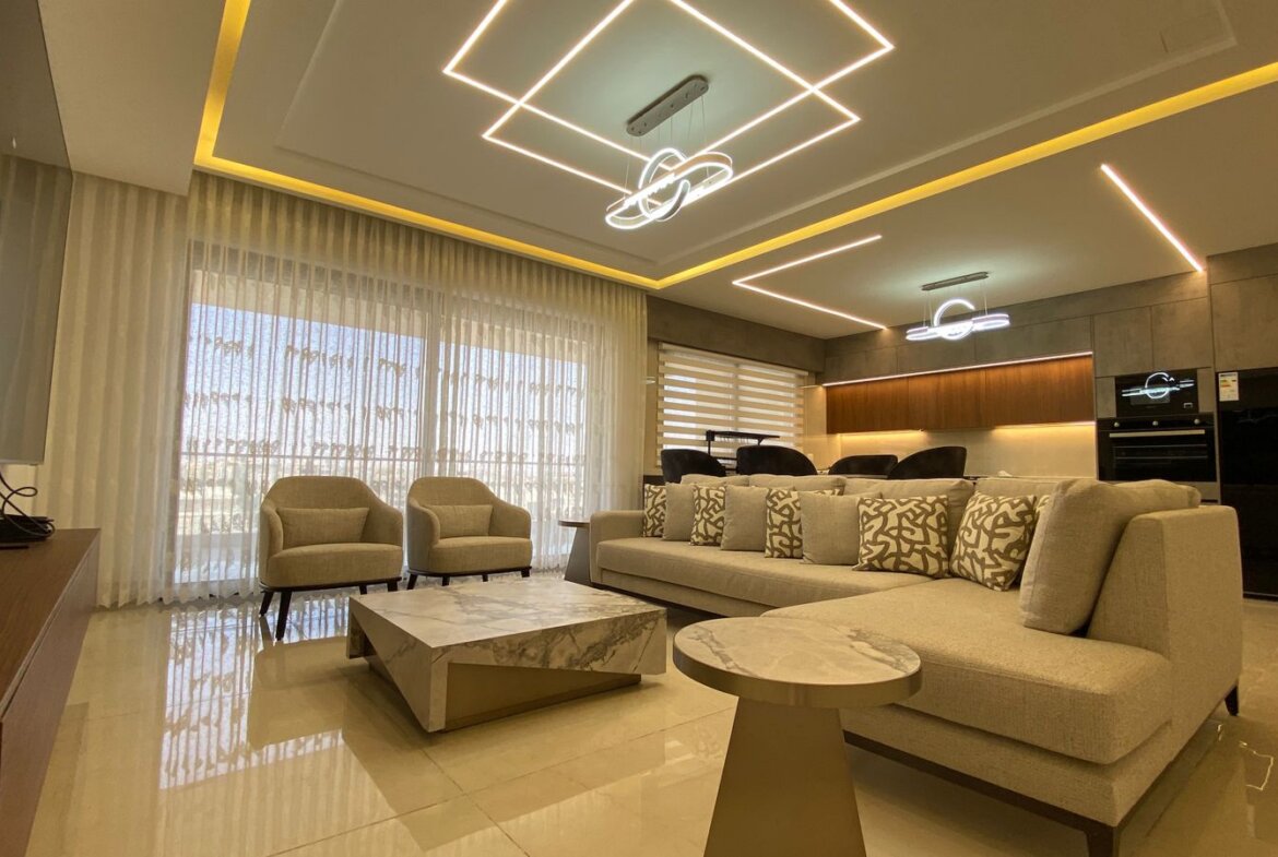 Luxurious living room with beige sectional sofa, patterned cushions, two beige armchairs, and marble coffee table facing a TV wall.