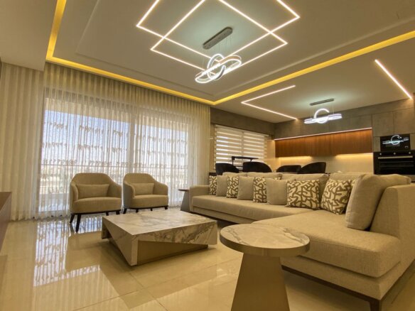 Luxurious living room with beige sectional sofa, patterned cushions, two beige armchairs, and marble coffee table facing a TV wall.