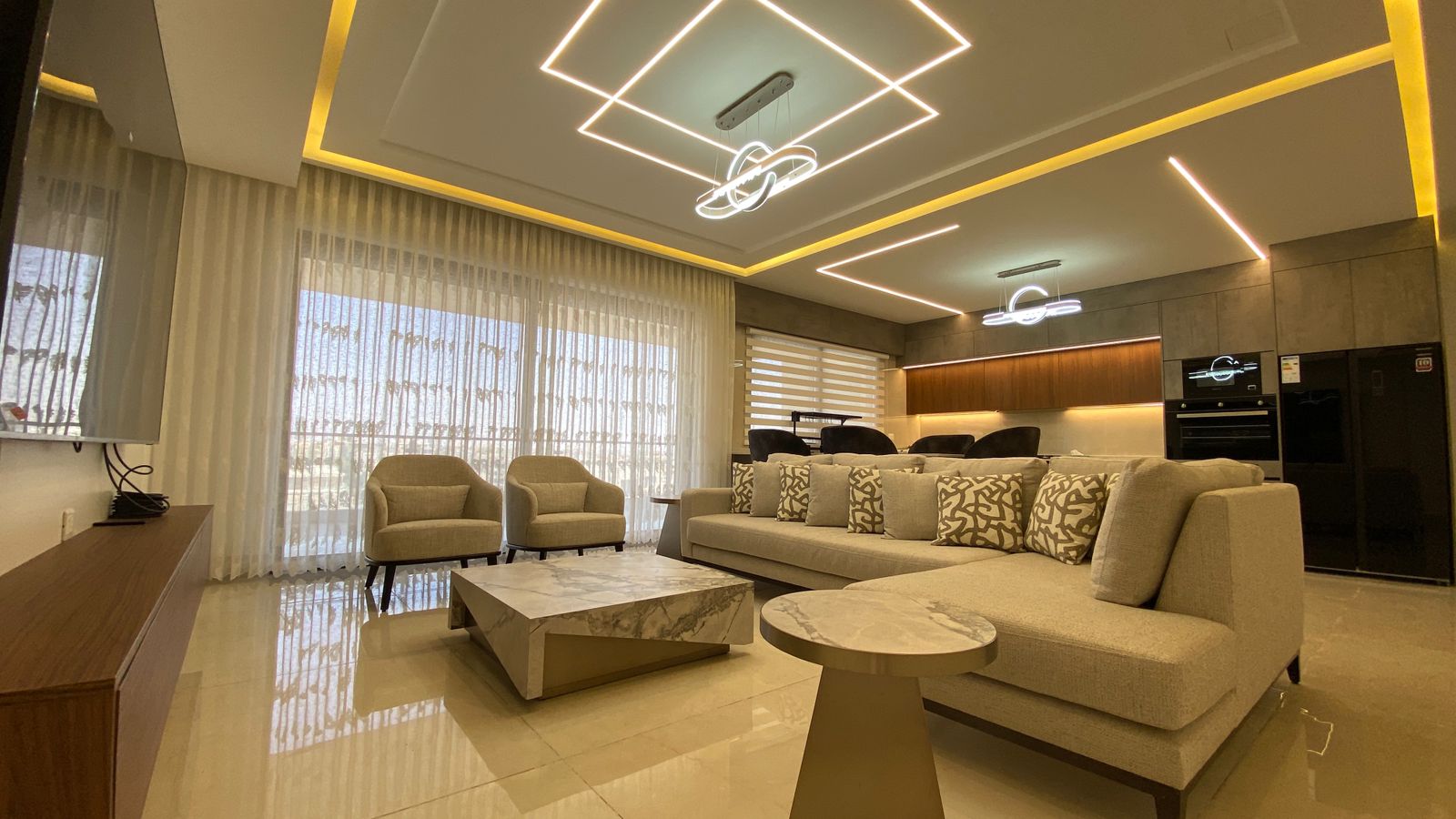 e3b44296-437d-4e12-ad83-8891aa2a7f25 Luxurious living room with beige sectional sofa, patterned cushions, two beige armchairs, and marble coffee table facing a TV wall.