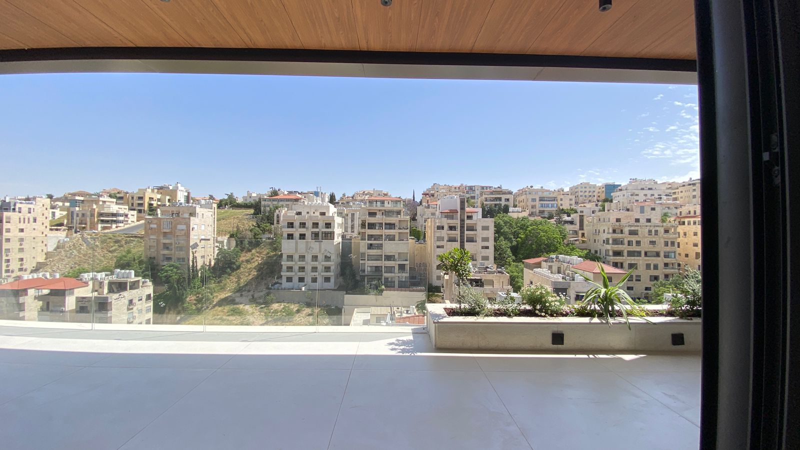 e3f84265-c35b-4d53-951a-31cf6cefe624 View of a hillside residential area seen from a modern balcony with a glass railing and planter boxes; blue sky above.