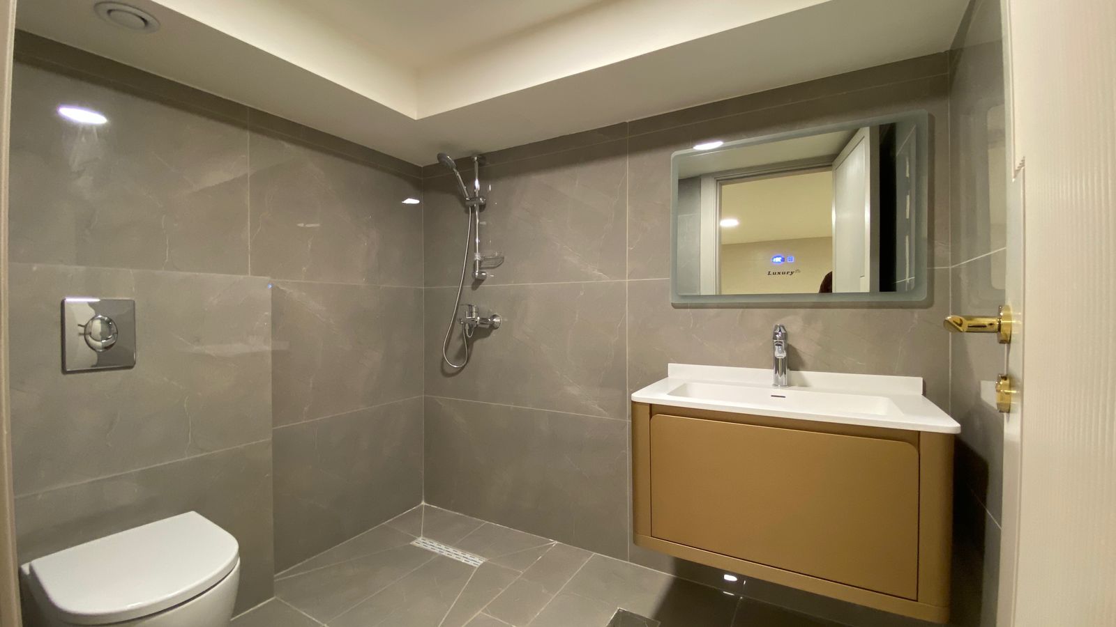 e3fd0acb-ef6e-44af-a1f5-838a07455589 Modern gray-tiled bathroom with a wall-mounted toilet, walk-in shower with handheld shower, and a beige vanity with white countertop and mirror.