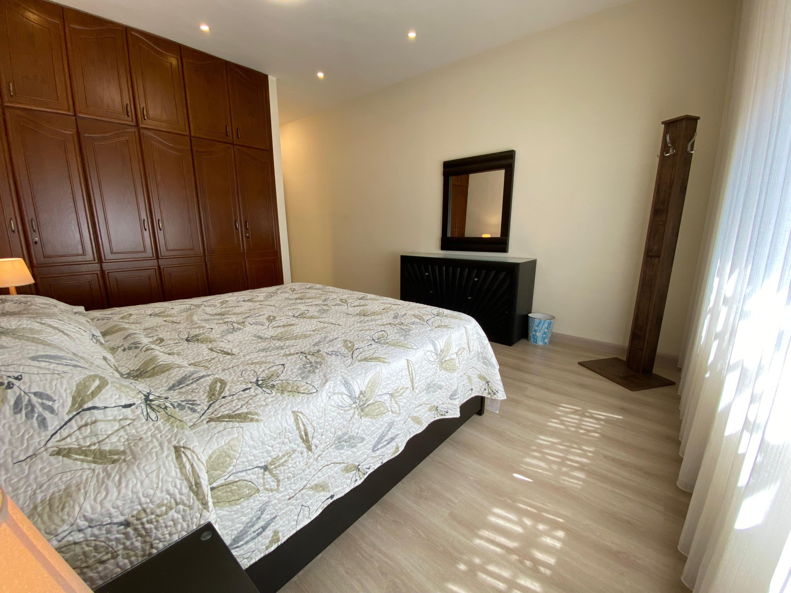 e45dce78-e1fb-4ec6-96e9-d203c3457a0f A bright bedroom with a large dark wood wardrobe along the left wall and a bed in the foreground with a leafy quilt pattern.