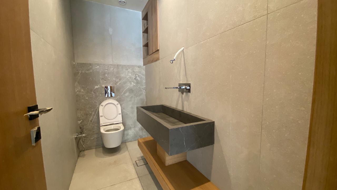 e47e9646-6ec3-40fe-be5a-d26299e46604 Narrow modern bathroom with a dark rectangular sink, wall faucet, and a white toilet at the far end. Gray marble walls and wooden shelf below the sink.