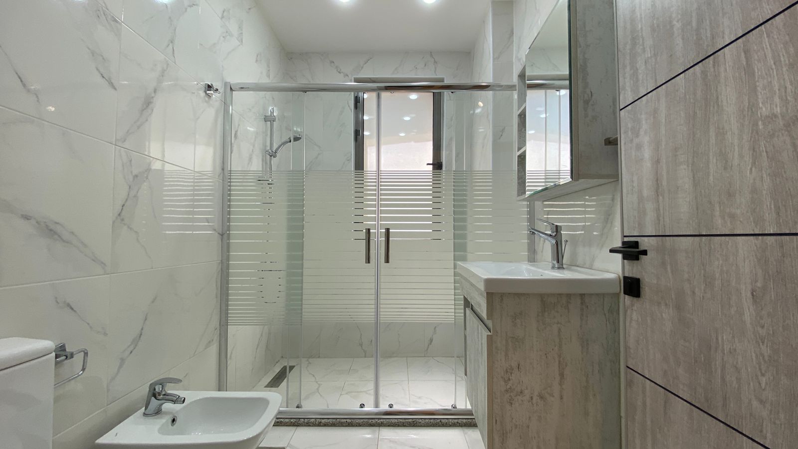e4fb49fc-5f78-4d2b-81ed-878580e44ae9 Modern bathroom with a glass shower enclosure, white sink vanity, and marble-look tiled walls and floor in a bright space.