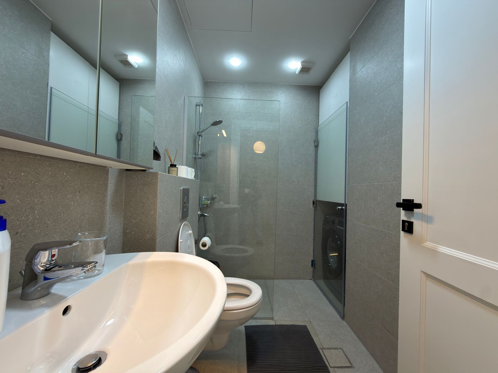 e547fc0a-86f8-40c2-b294-674a1ceeaa9b Modern grey-tiled bathroom with a glass-enclosed shower, rainfall showerhead, and a white porcelain sink in the foreground.