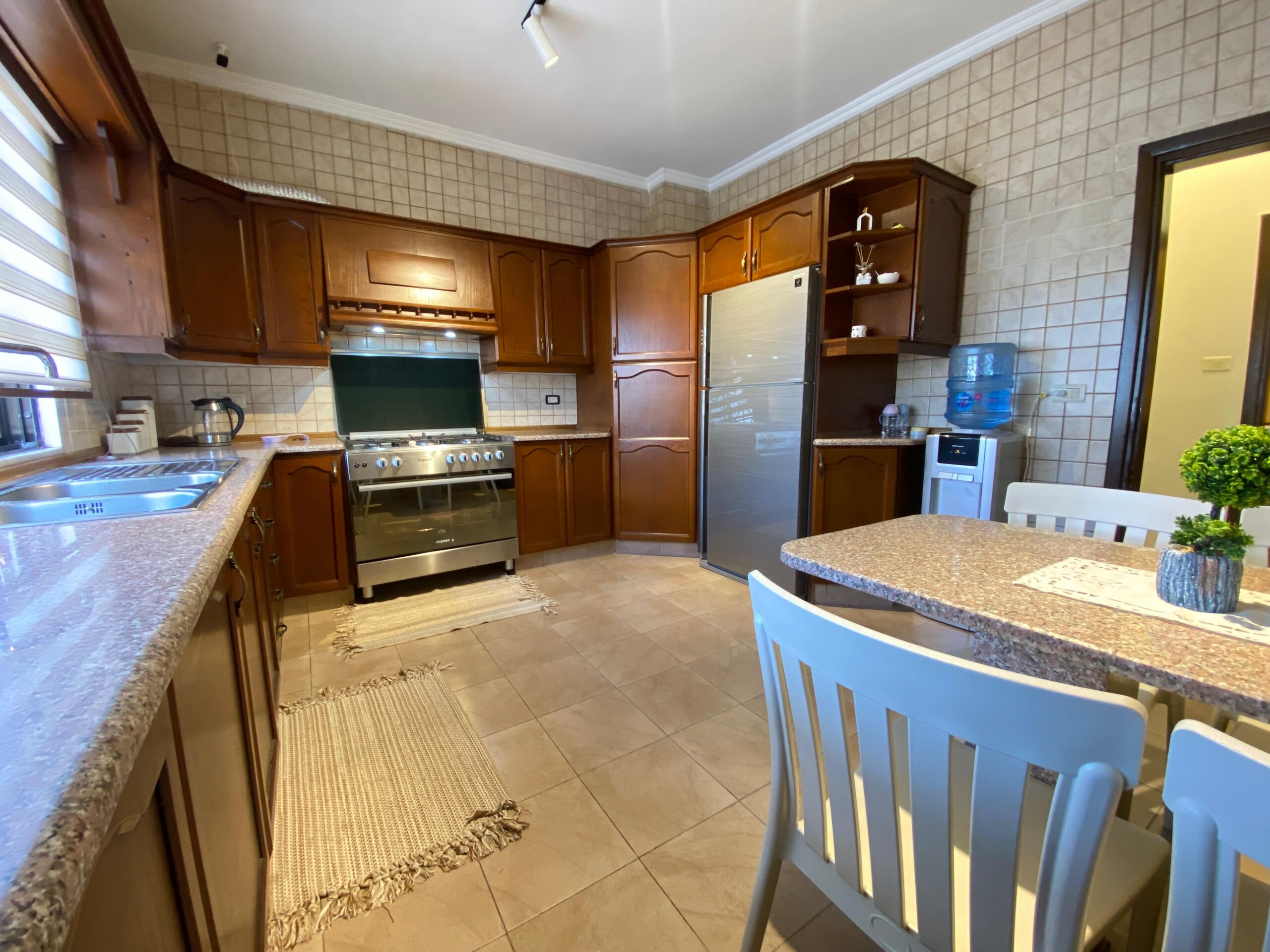 e5a0e58b-50a8-45eb-b6cb-72c7b52a46a3 Spacious kitchen with dark wood cabinets, stainless steel stove, and a blue-dining set around a granite island.
