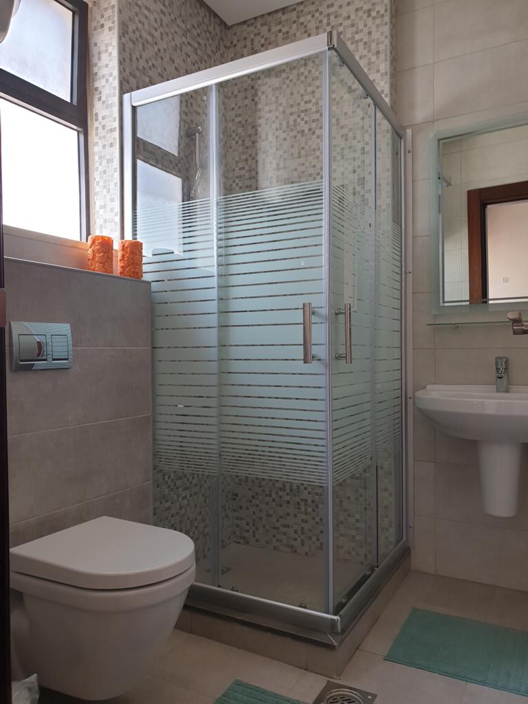 e5b4c269-f793-4f37-a9ec-3b8c2c8b92c1 Modern bathroom with a glass-framed shower enclosure featuring frosted stripes, a wall-mounted toilet, and a pedestal sink nearby.