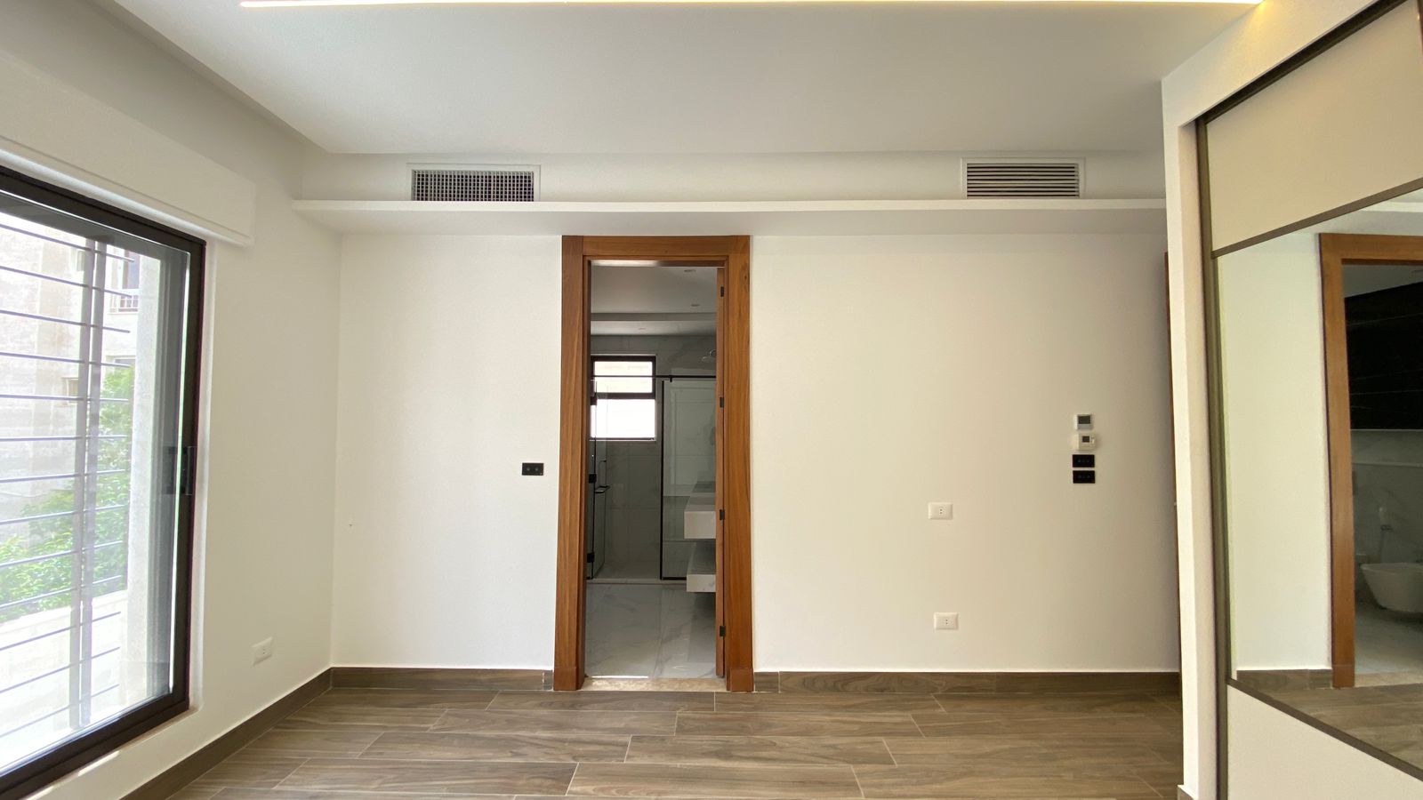 e6247746-dcbc-41bf-beb4-e1f34c1d2040 Empty modern room with a wood-framed doorway leading to a glass-enclosed bathroom, left a large window, and right a mirrored closet door.