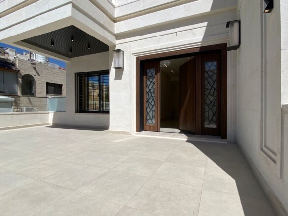Open double wooden doors with decorative glass panels reveal a lit interior, on a large sunlit tiled terrace of a modern white building.